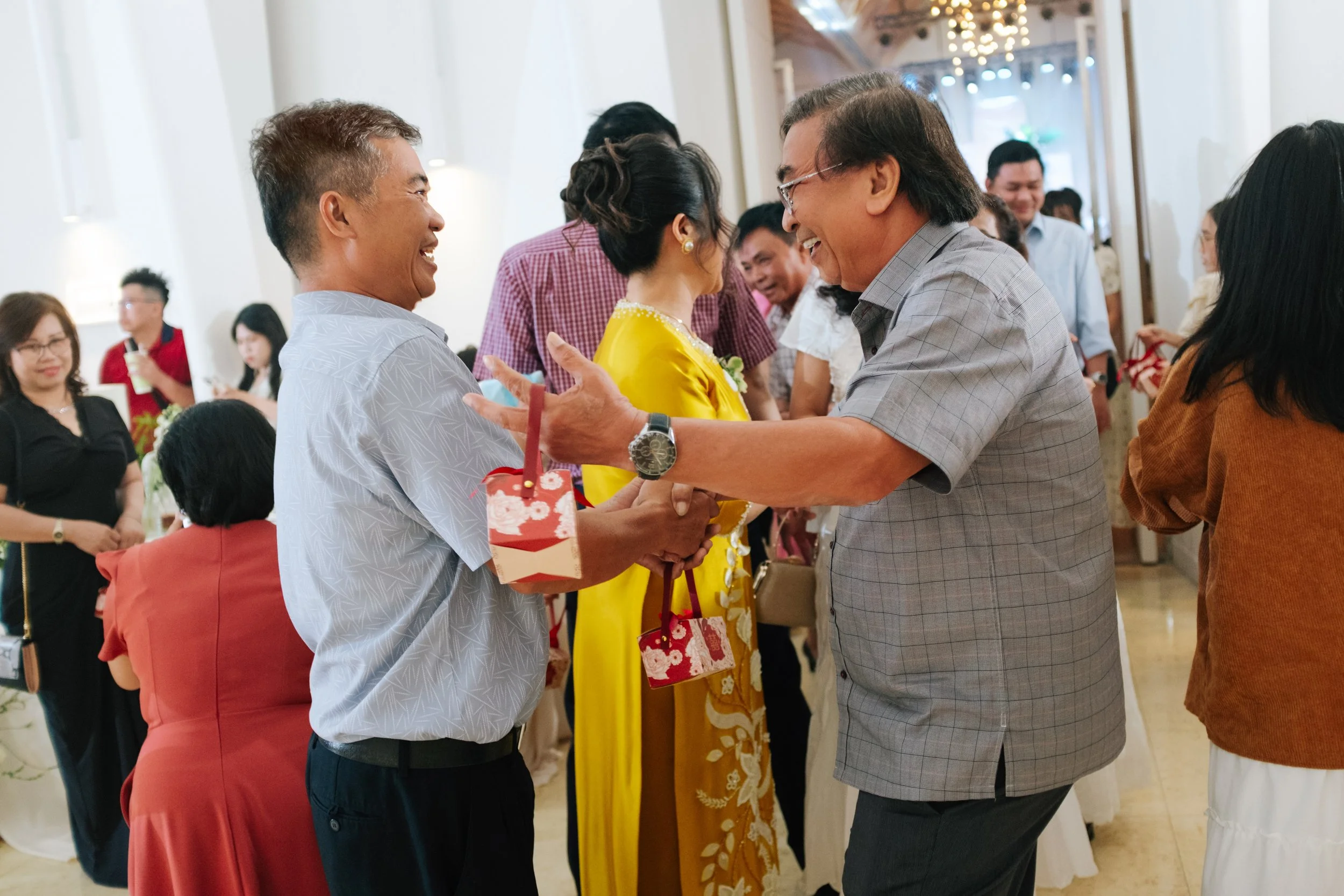 Two men smiling and shaking hands at a social gathering, with a woman in a yellow dress in the background, surrounded by other people in a well-lit indoor setting.