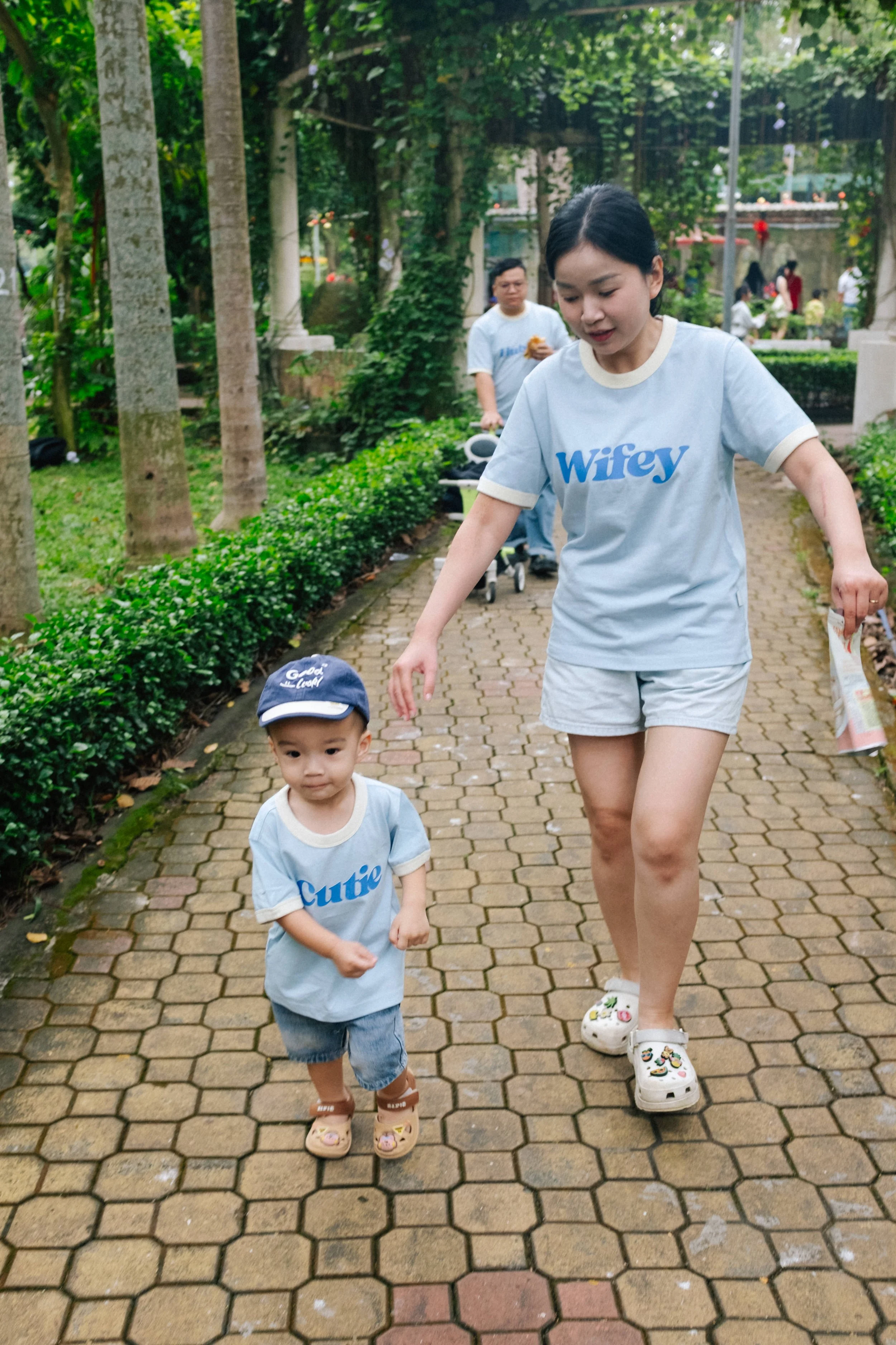 A woman and a young boy walking on a brick path in a park with green trees and bushes. The woman is wearing a light blue shirt reading 'Wifey' and shorts, and the boy is wearing a matching light blue shirt reading 'Cute,' shorts, a cap, and Crocs.