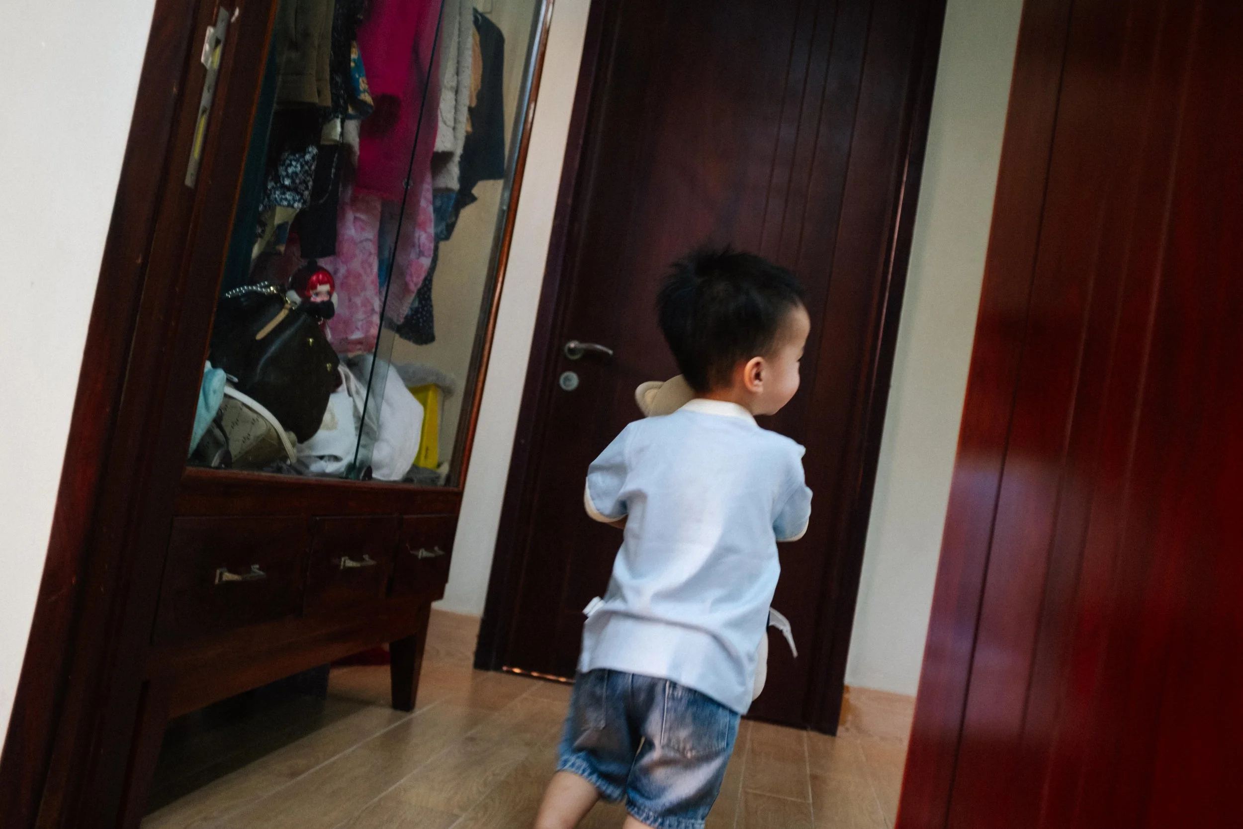 A young boy in a light blue shirt and denim shorts running inside a room with wooden furniture and a wardrobe with clothes inside.
