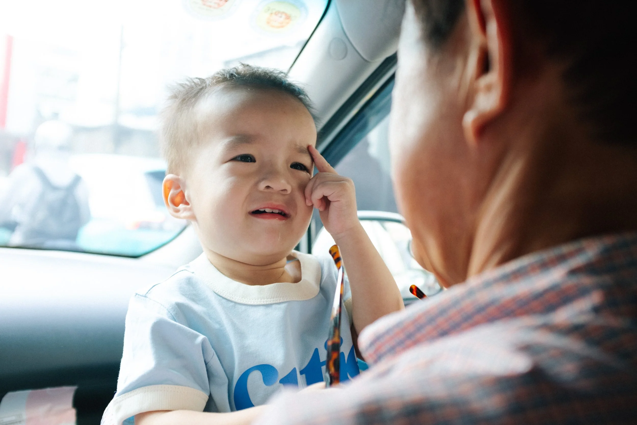 A young child with a sad expression, touching their eyebrow, looking at an older person inside a car.