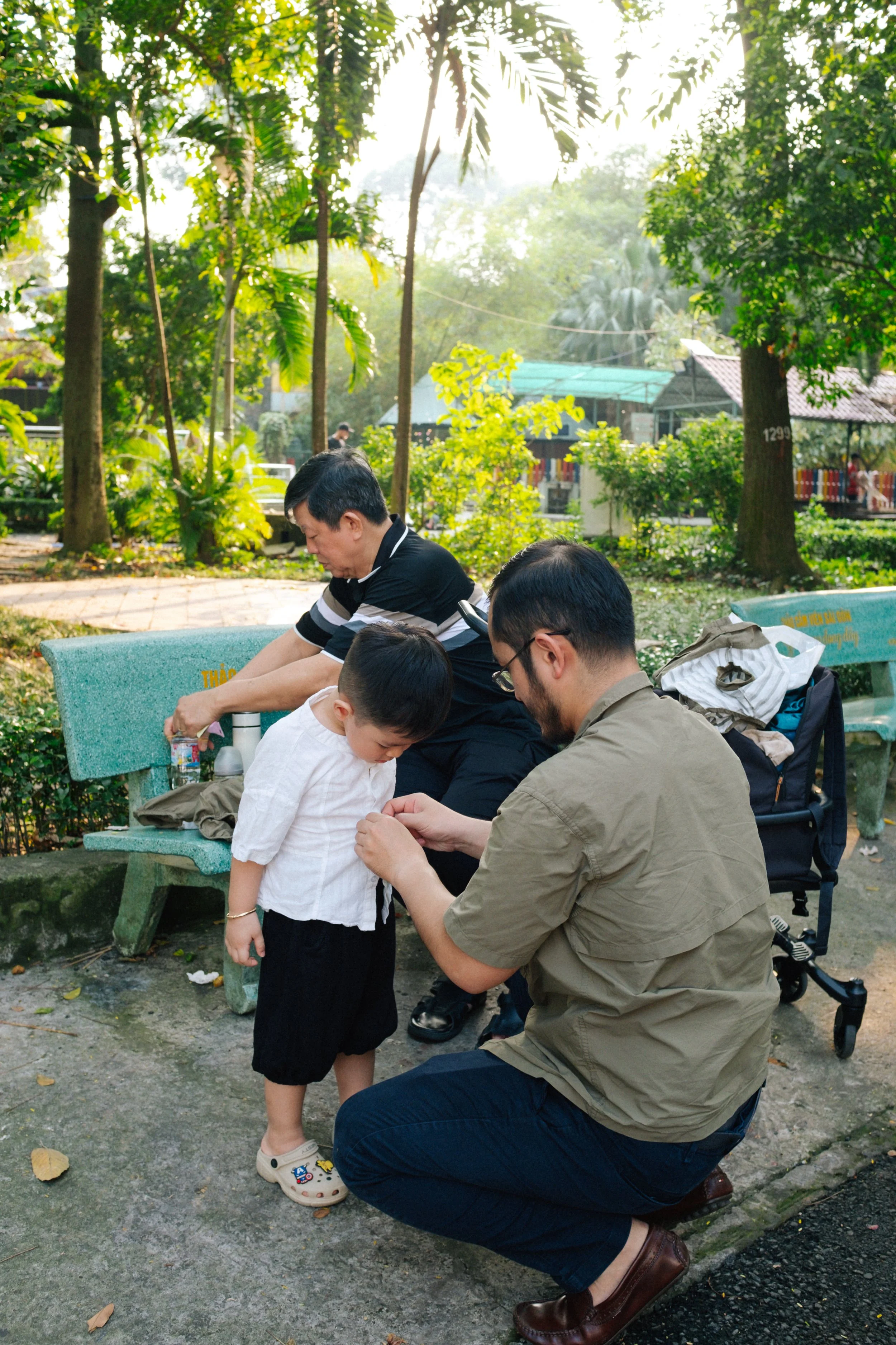A young boy standing while an adult helps him put on a bracelet in a park with trees and benches.