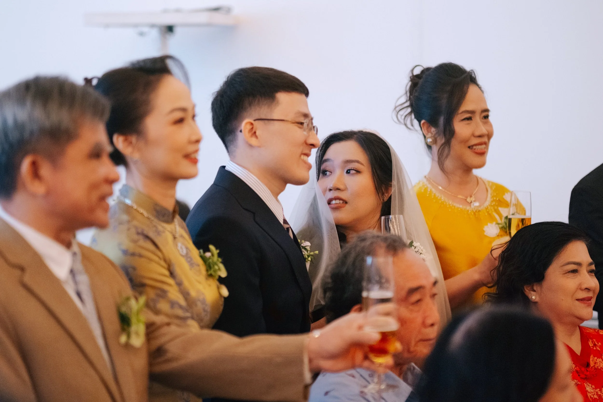 Wedding celebration with multiple guests, including a man in a black suit and a woman in a wedding dress, standing close together and holding drinks.