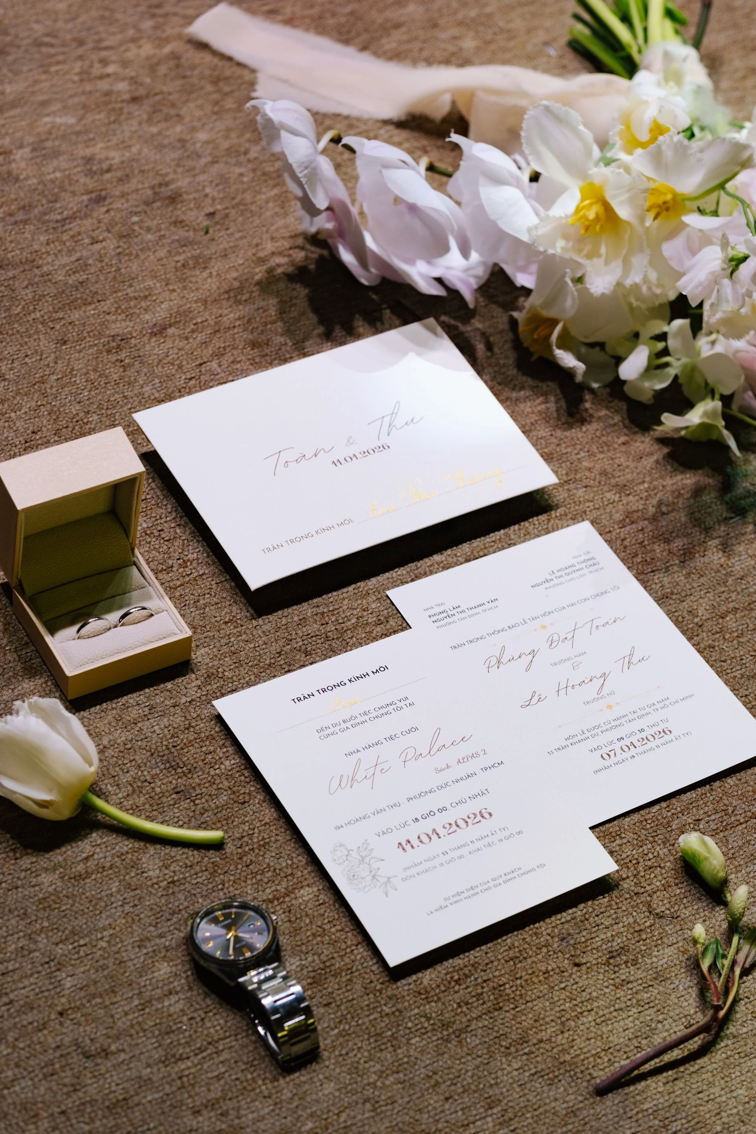 Wedding invitations, rings in a box, a watch, and flowers on a brown carpet.