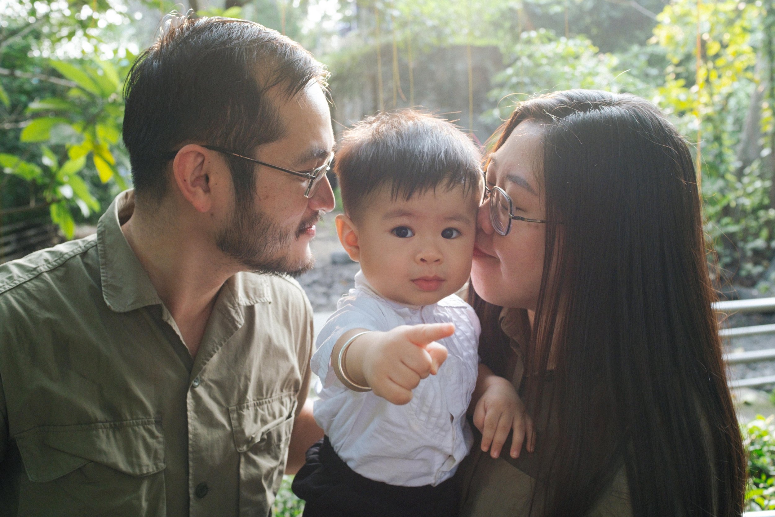 A couple with a young child outdoors surrounded by greenery. The woman is kissing the child's cheek while holding him, and the man is smiling, looking at them.