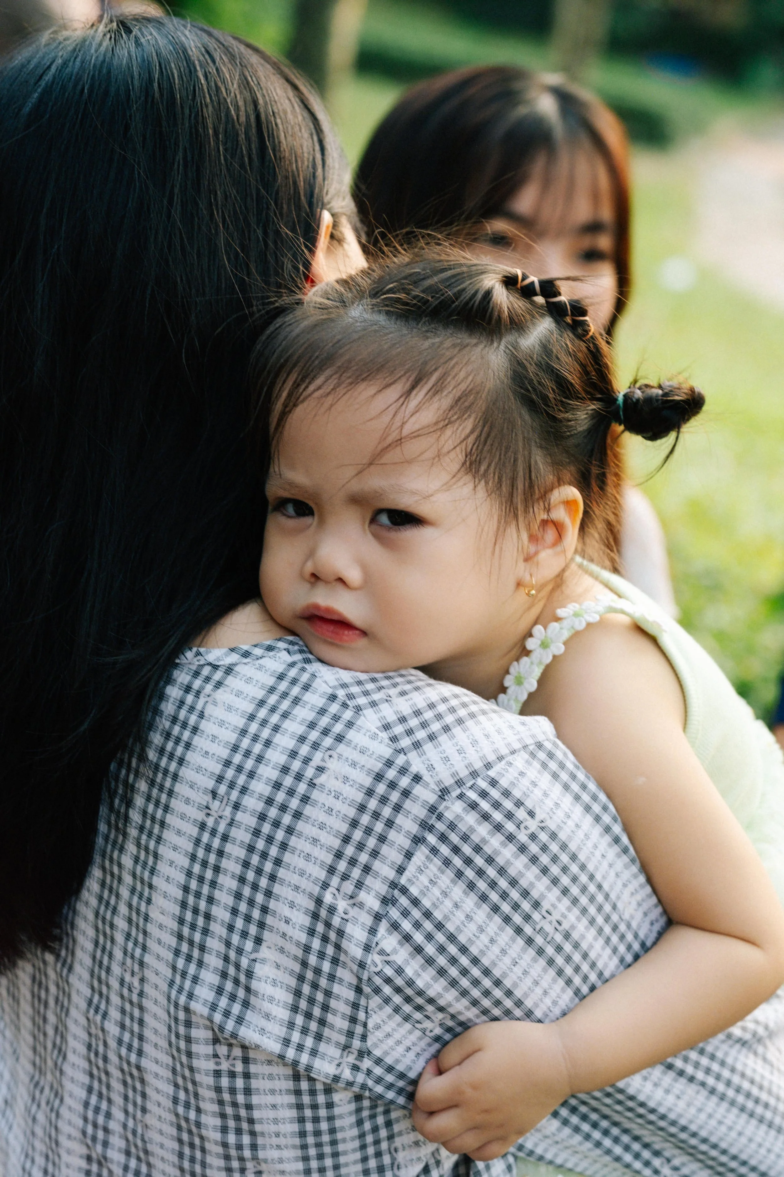 A young girl with a serious expression hugging an adult, with another adult in the background outdoors.