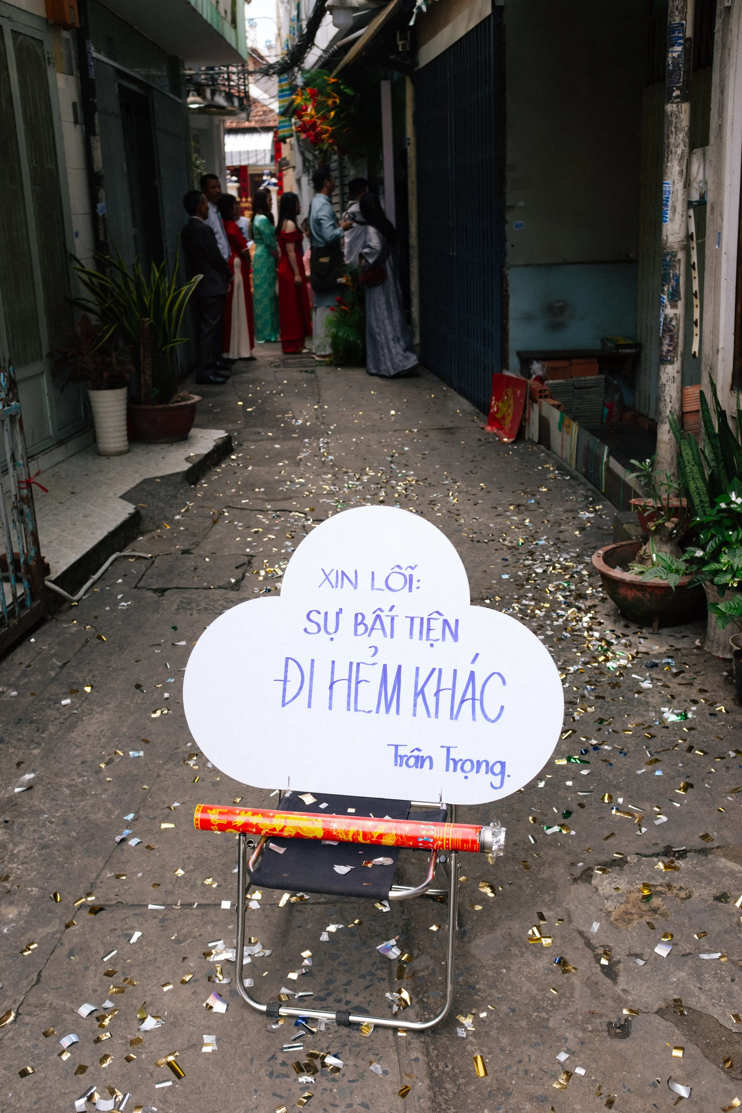 A street scene with a sign on a chair that reads, "Sorry for the inconvenience, see you another day, Trần Trọng," in Vietnamese. Confetti is scattered on the ground, and people dressed in traditional and formal attire are gathered further down the al