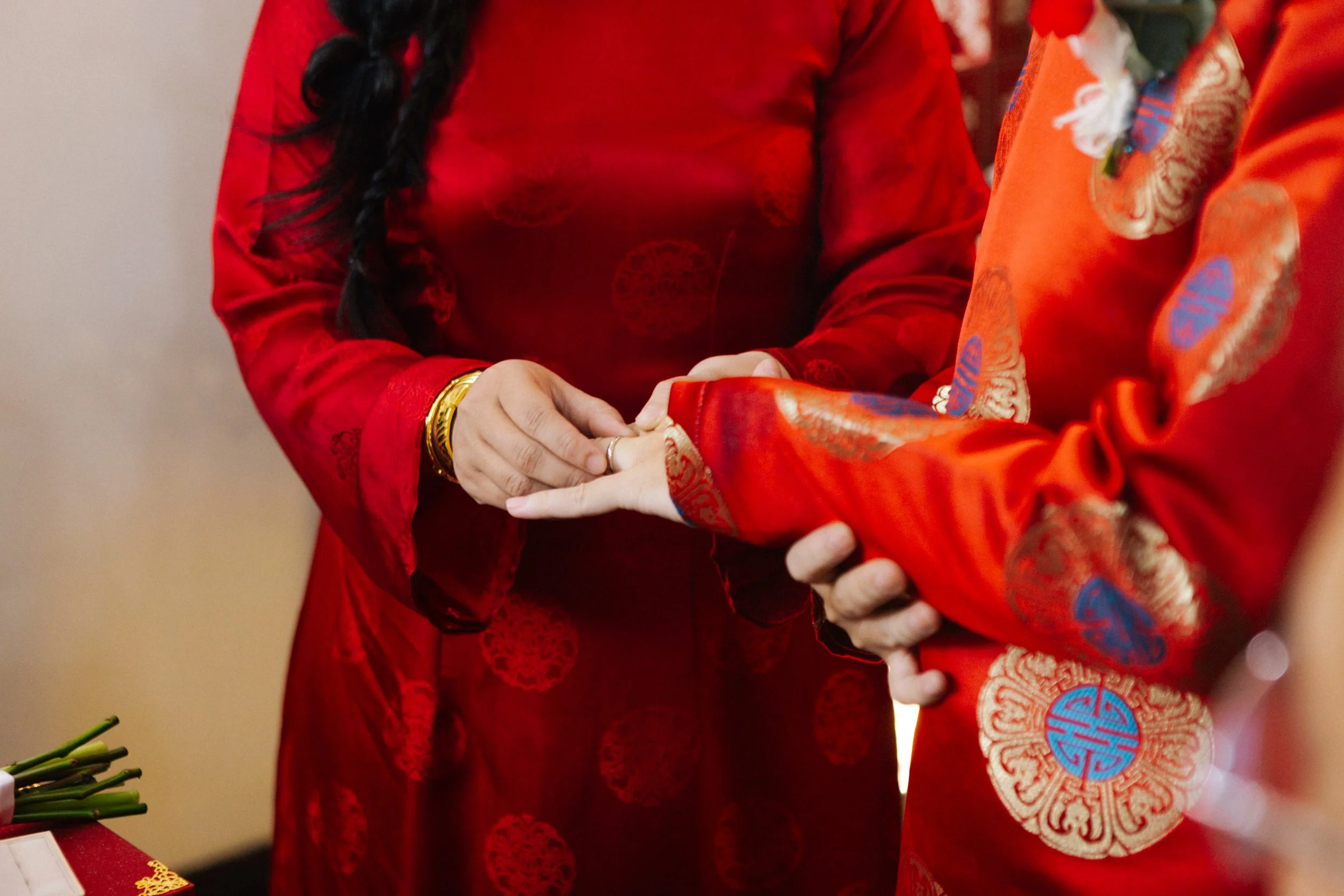 Close-up of a couple holding hands during a traditional wedding ceremony, with couples wearing red attire with gold and blue embroidery.