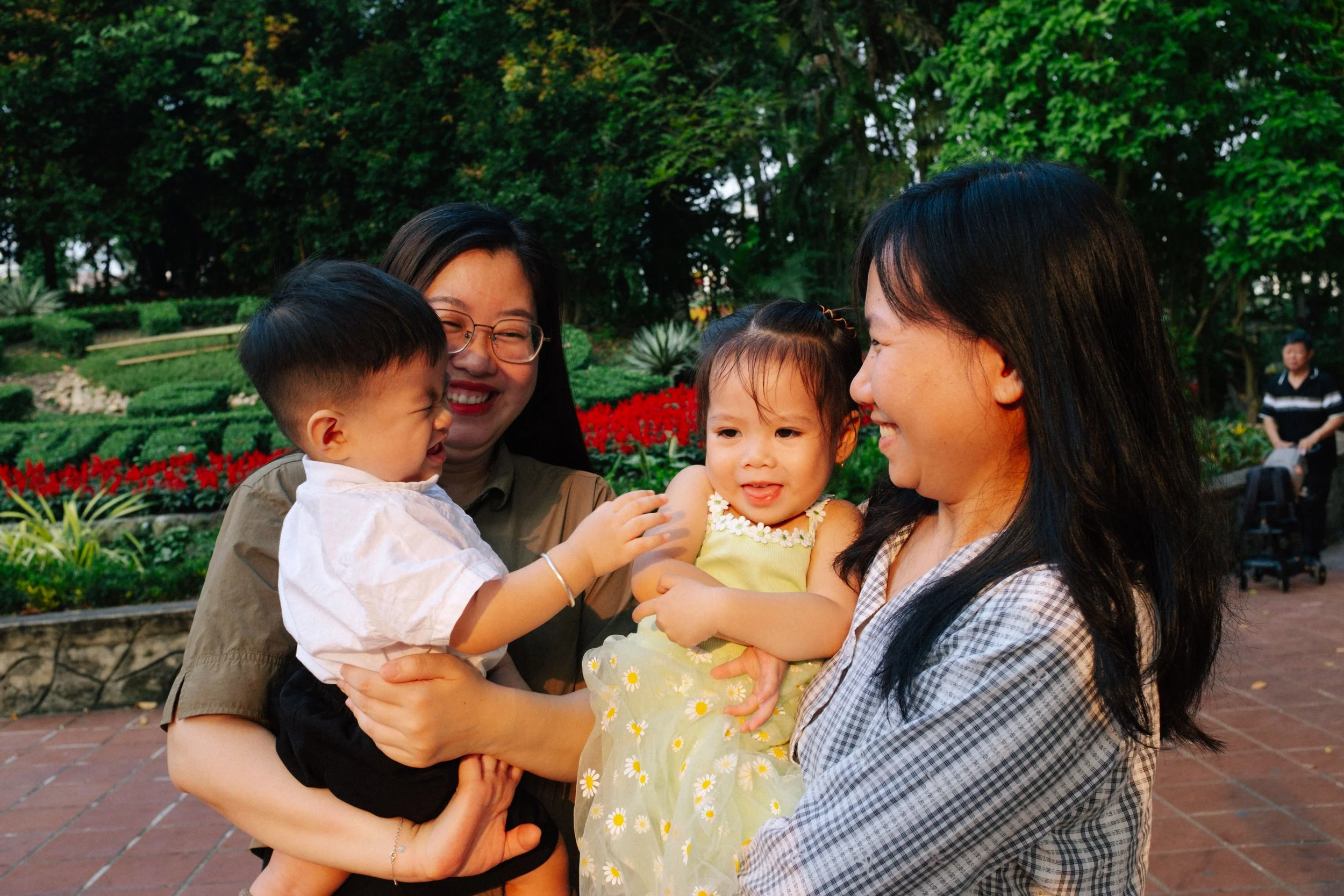 Two women holding two young children outdoors in a park with green trees and red flowers in the background.