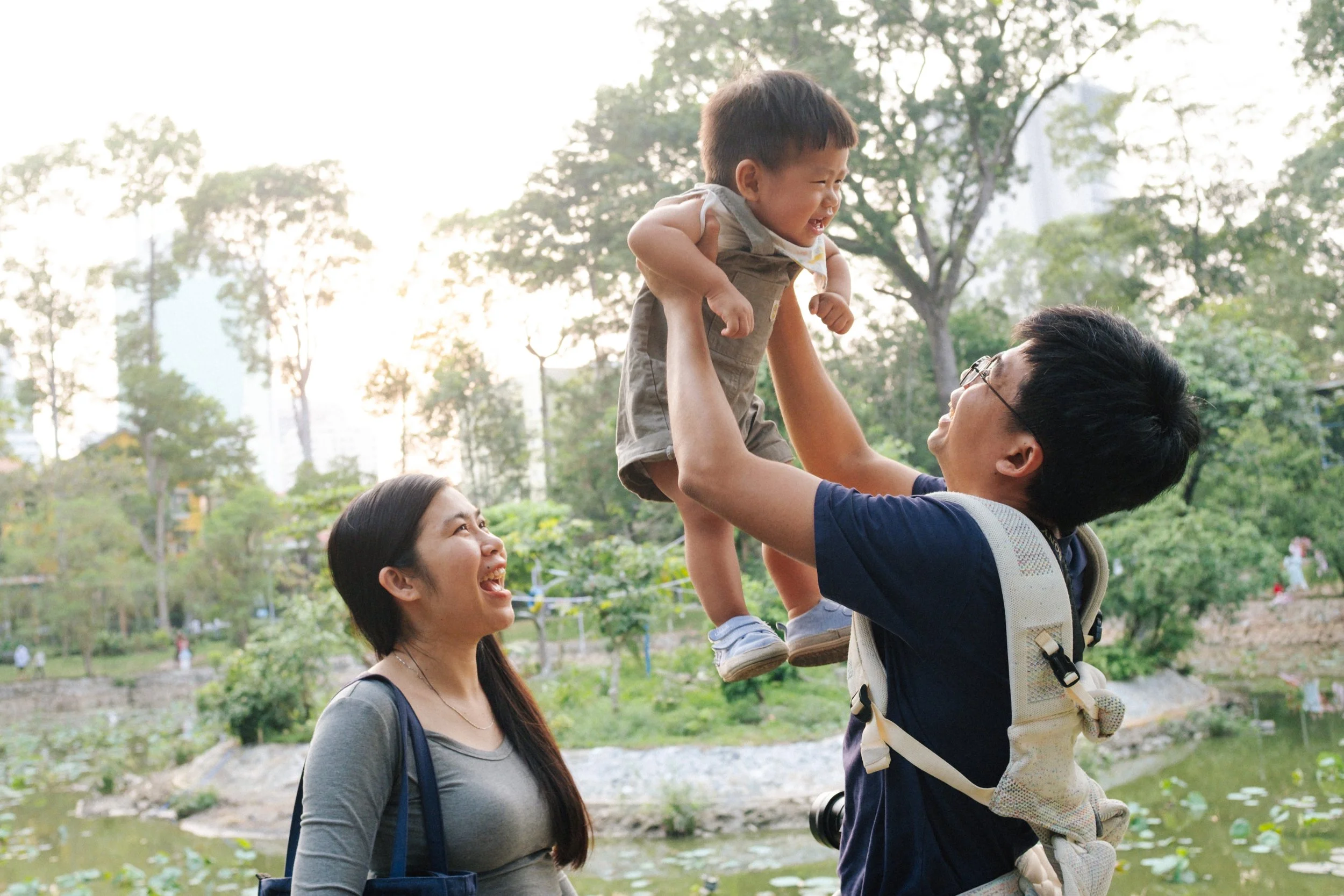 A man lifting a young boy in the air while a woman looks on smiling in a park with greenery and trees in the background.