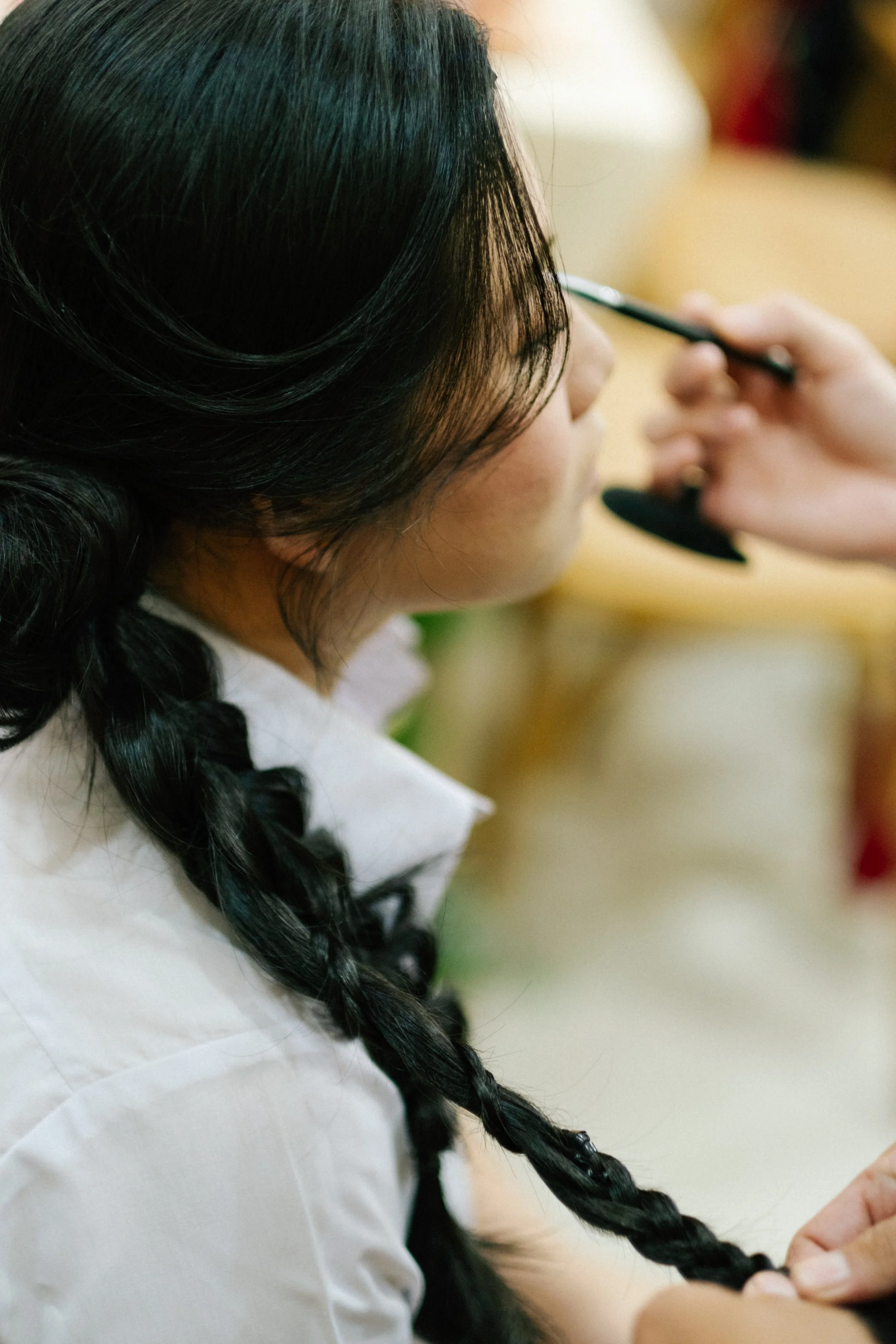 A woman with black braided hair is getting her makeup done by hand holding a brush near her face.