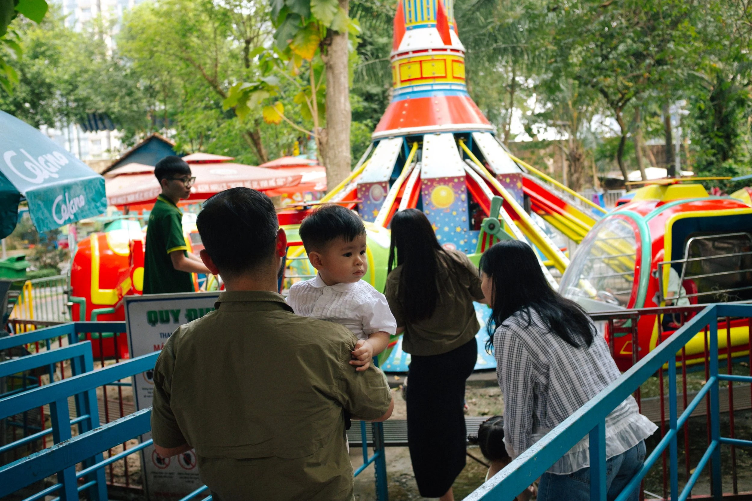People standing in line at a colorful amusement park ride, with some children and adults present, surrounded by trees and park stalls in the background.