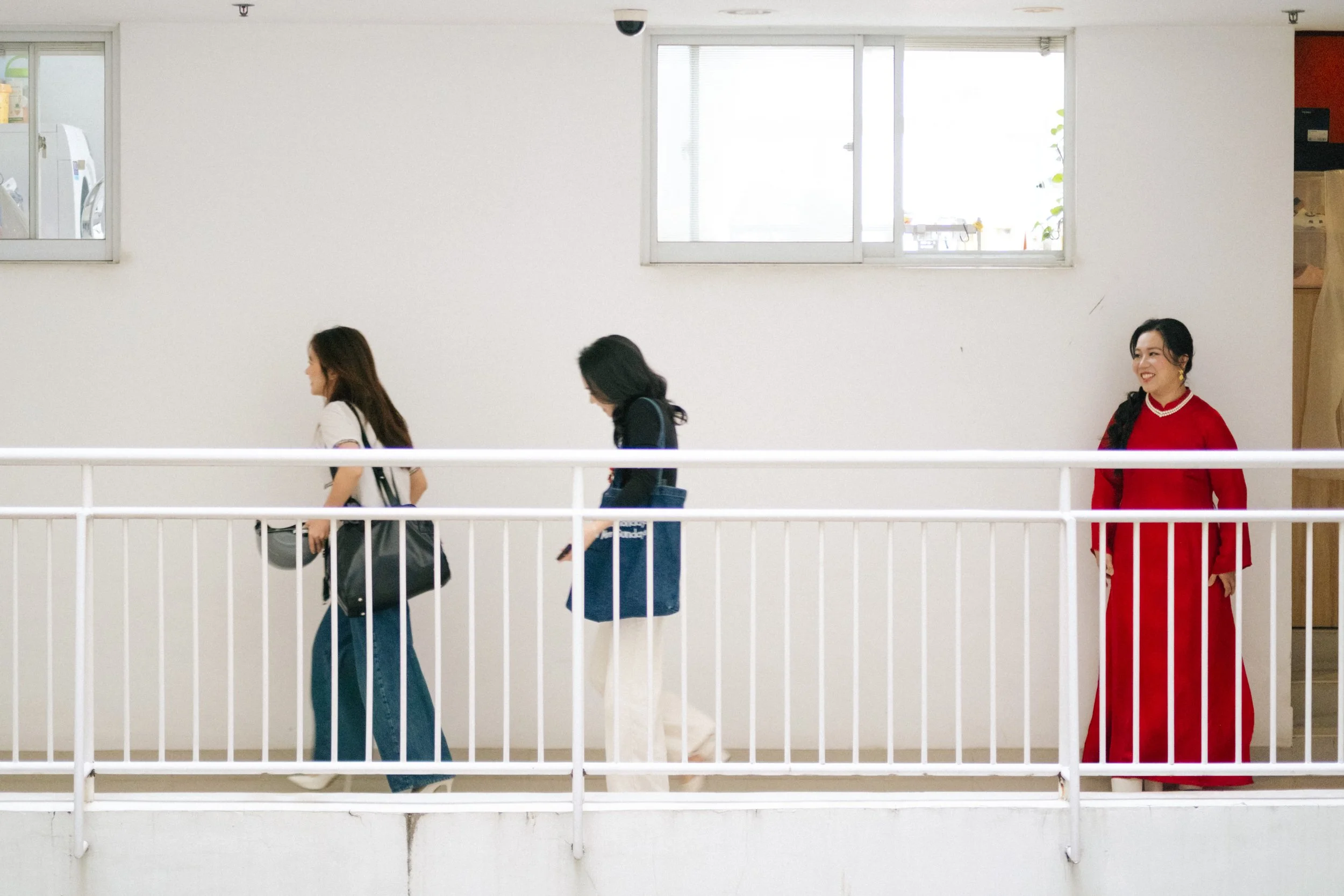 Three women standing on a white balcony with white railing, indoors, with large windows in the background. One woman in a red dress smiling at the third woman, who is wearing a black jacket and white pants. The woman in the middle is dressed casually