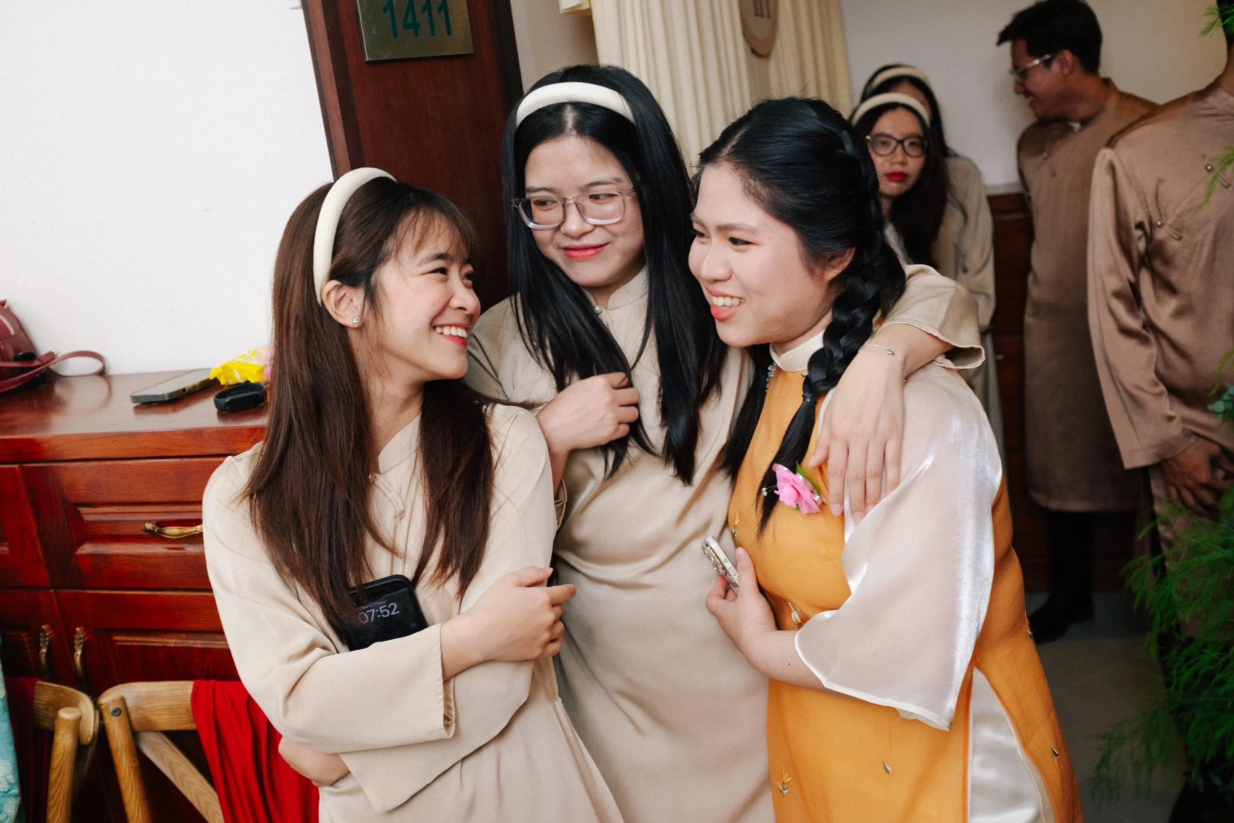 Three women wearing traditional Vietnamese áo dài, smiling and embracing each other at a gathering, with two more people in the background.