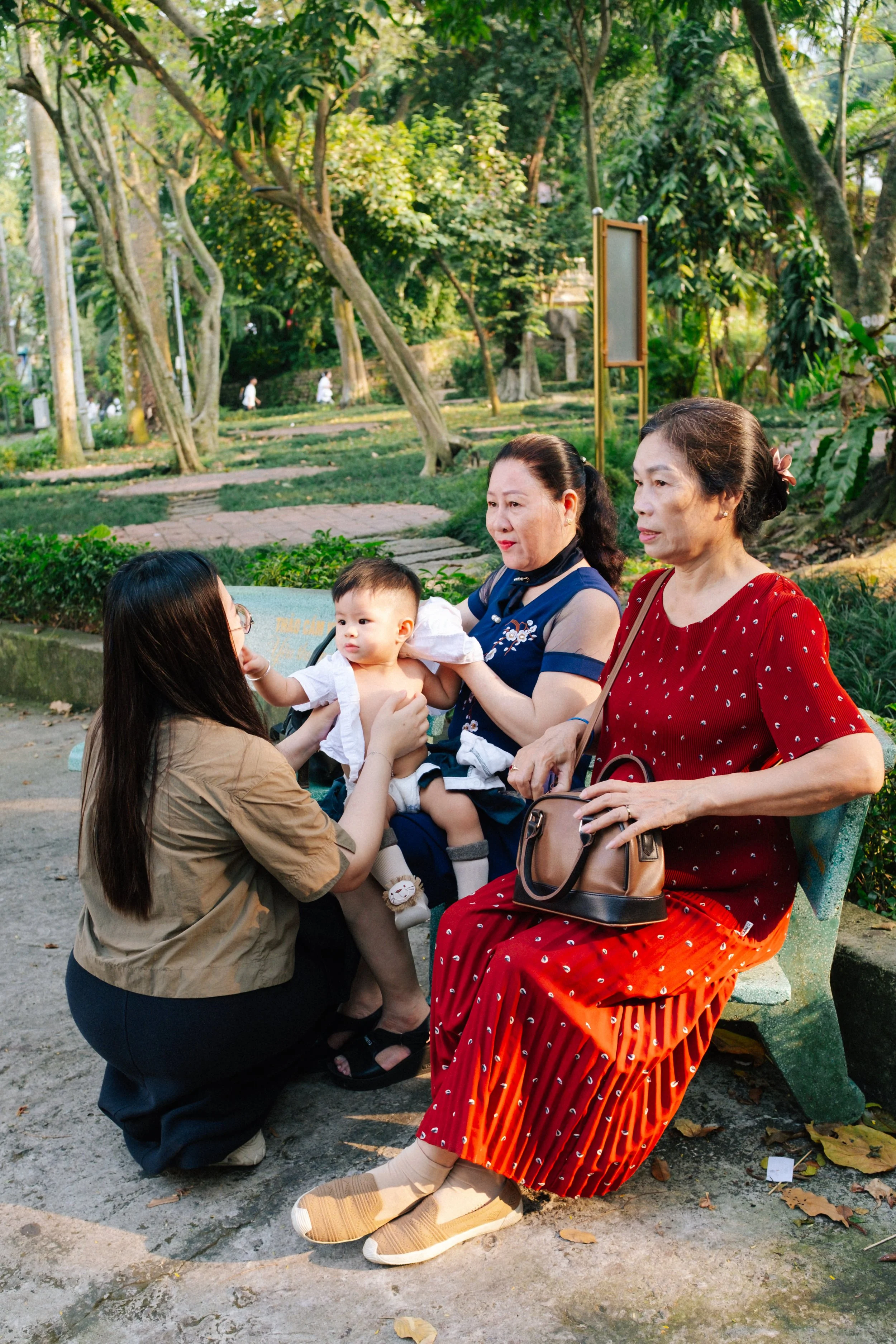 Three women and a baby sitting on a park bench, with one woman helping the baby to drink from a bottle. The park has tall trees and lush greenery in the background.
