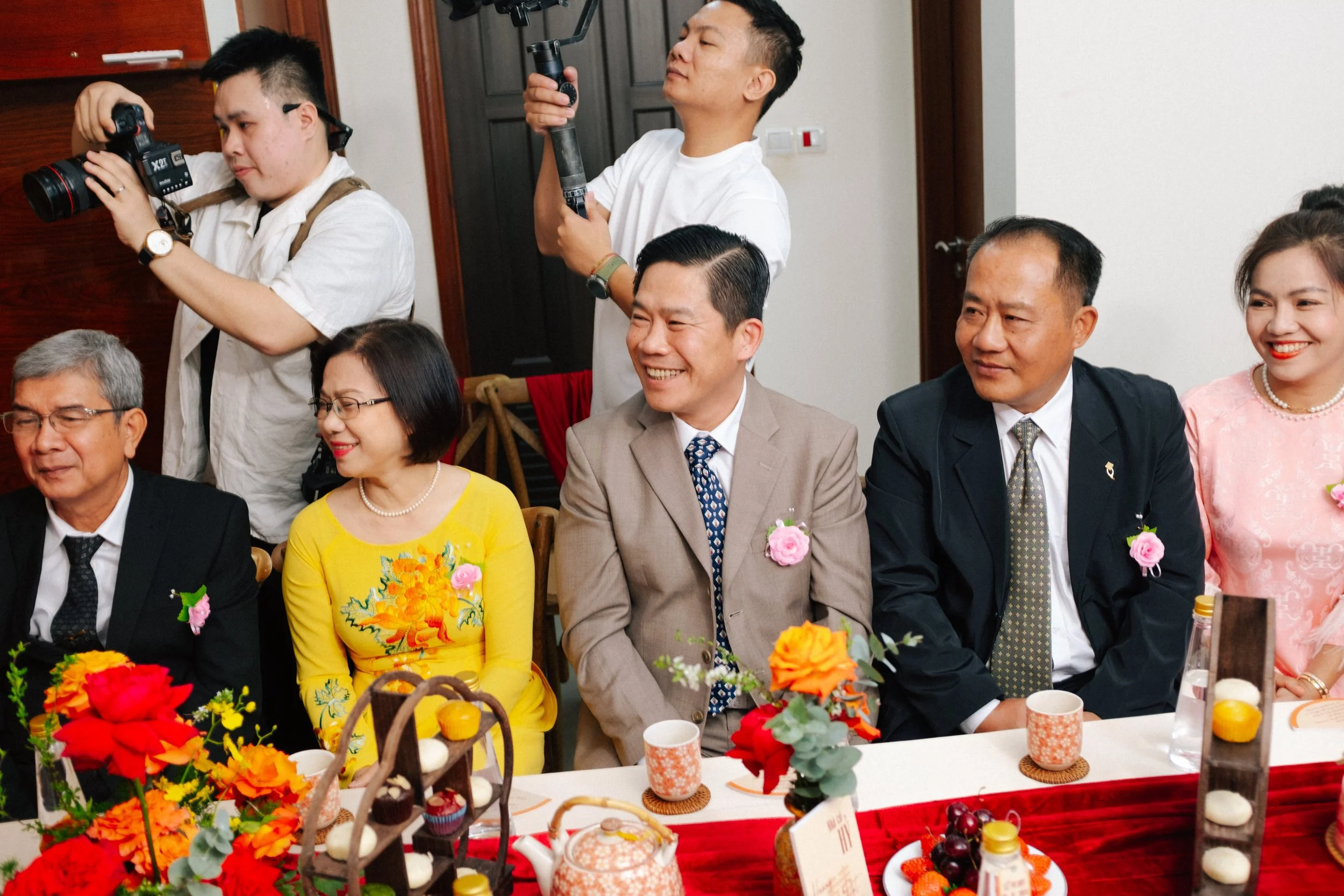 People sitting at a decorated table during a formal event, with cameras recording in the background.