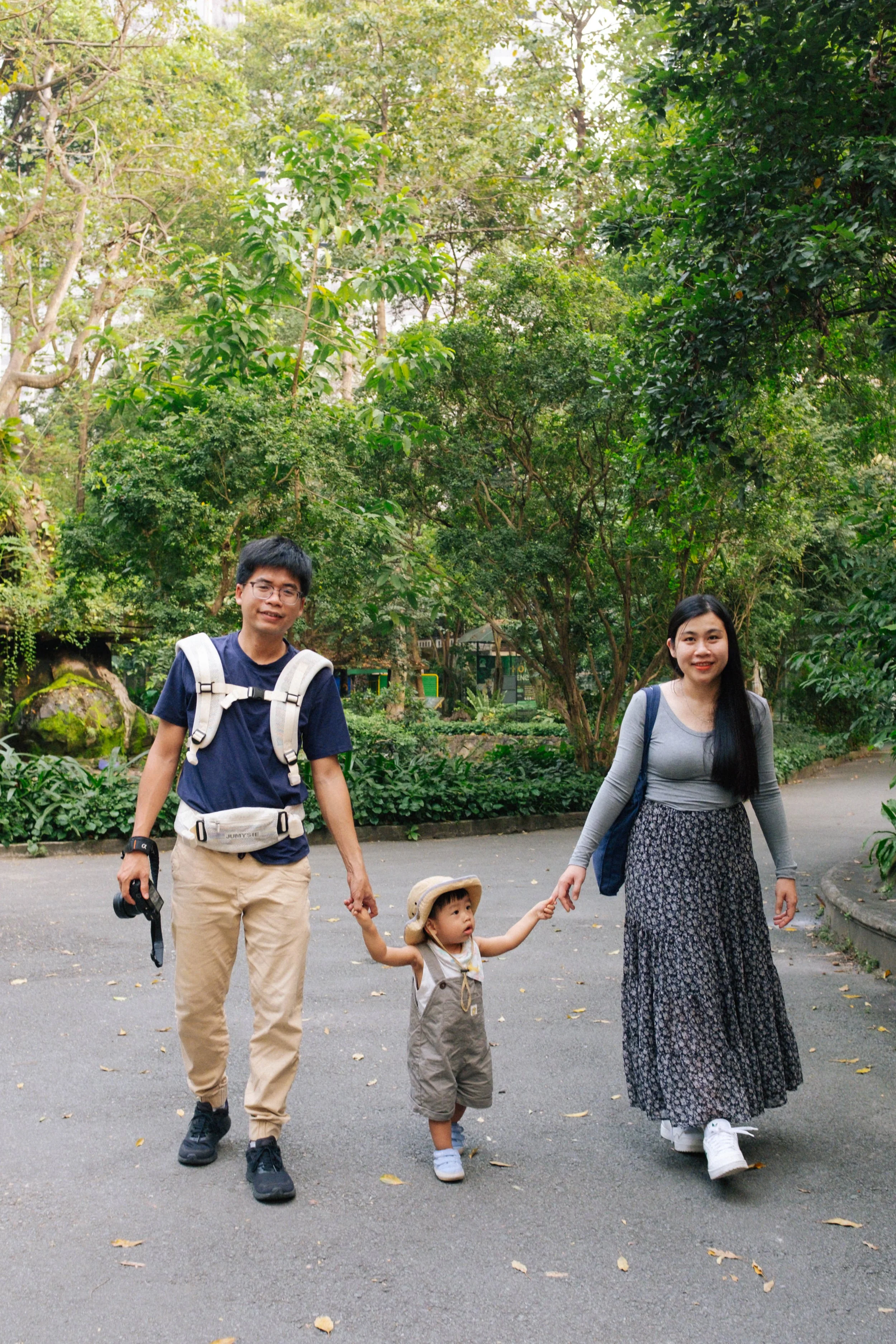 A family of three walking in a green park, holding hands. The dad is on the left wearing glasses, a navy shirt, and khaki pants. The young girl in the middle wears a hat and khaki overalls. The mom on the right has long black hair and wears a gray to