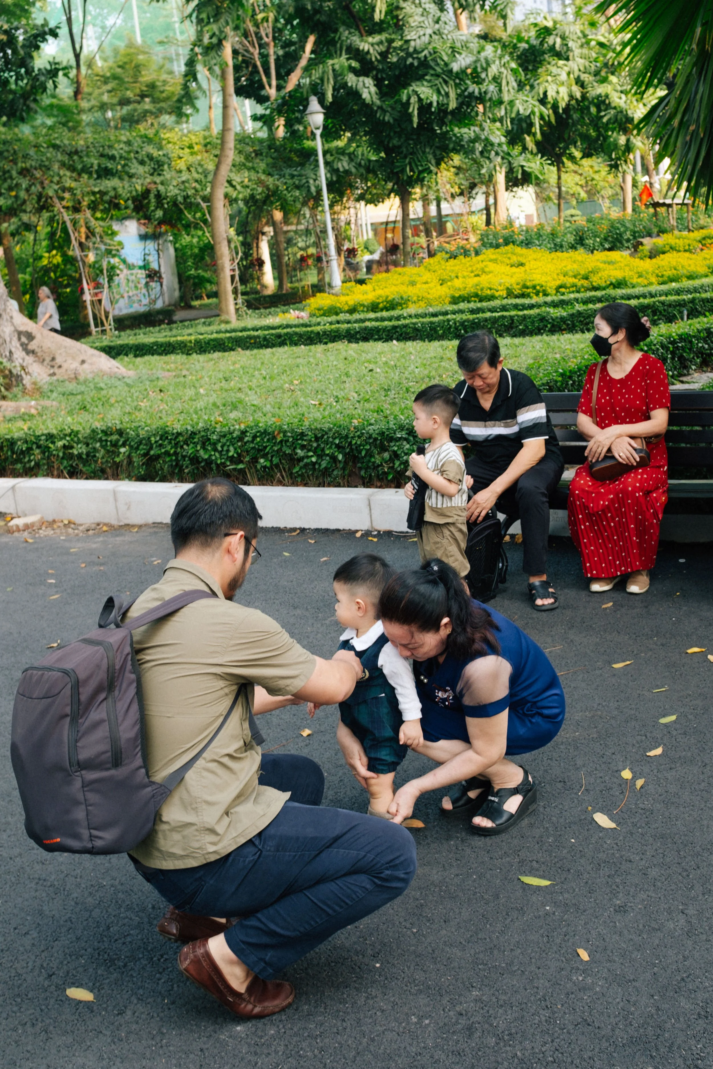 Family of five at a park, with two children and three adults, one woman crouching to help a young boy, and everyone else sitting on a bench or standing nearby.