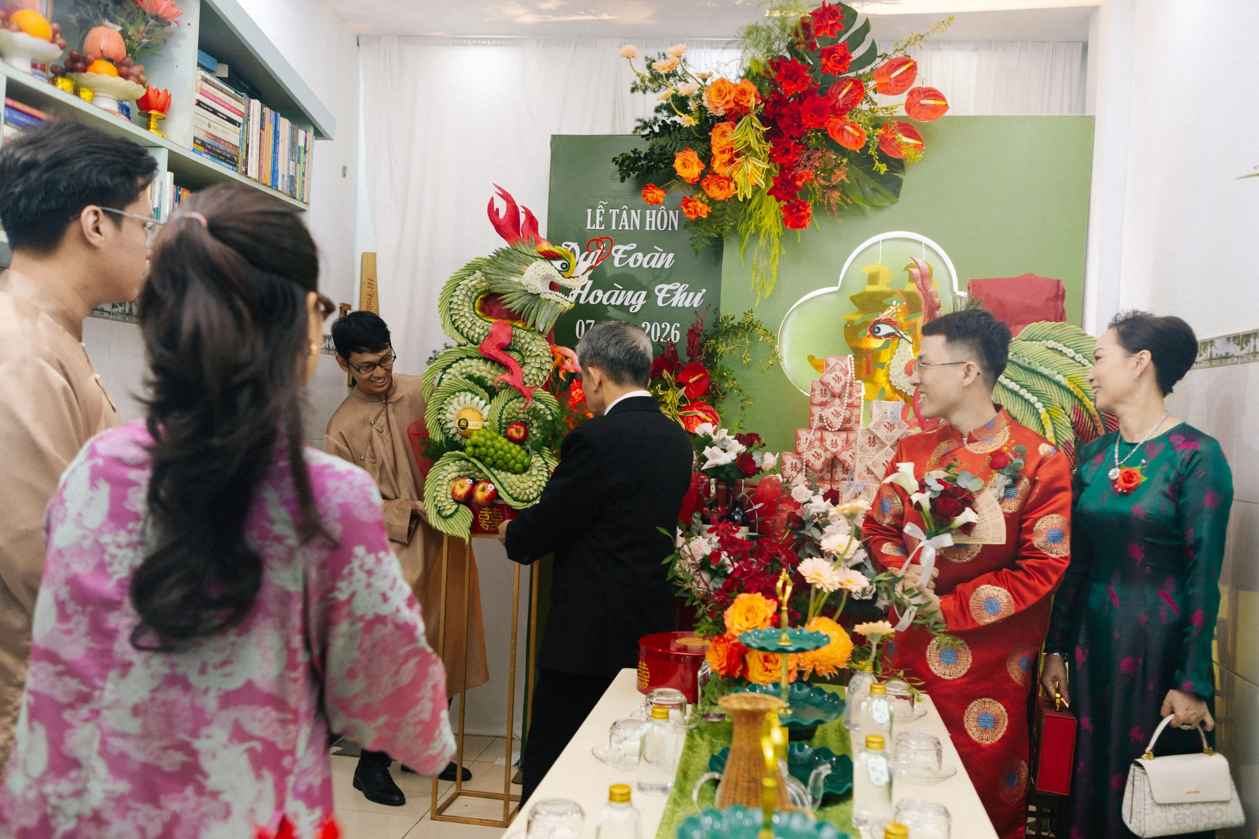 People celebrating a wedding with traditional Vietnamese decorations, including a dragon made of fruits and flowers, and a large floral arrangement with a green backdrop.
