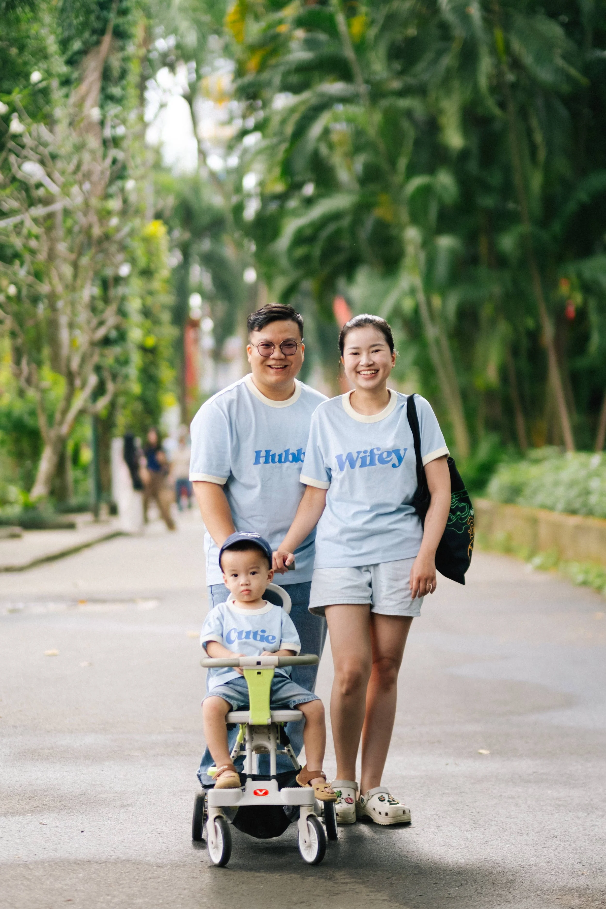 A family of three walking outdoors on a paved path surrounded by greenery. The father wears glasses and a shirt that says "Hubby," the mother wears a shirt that says "Wifey," and their young child sits in a stroller wearing a shirt that says "Cutie."