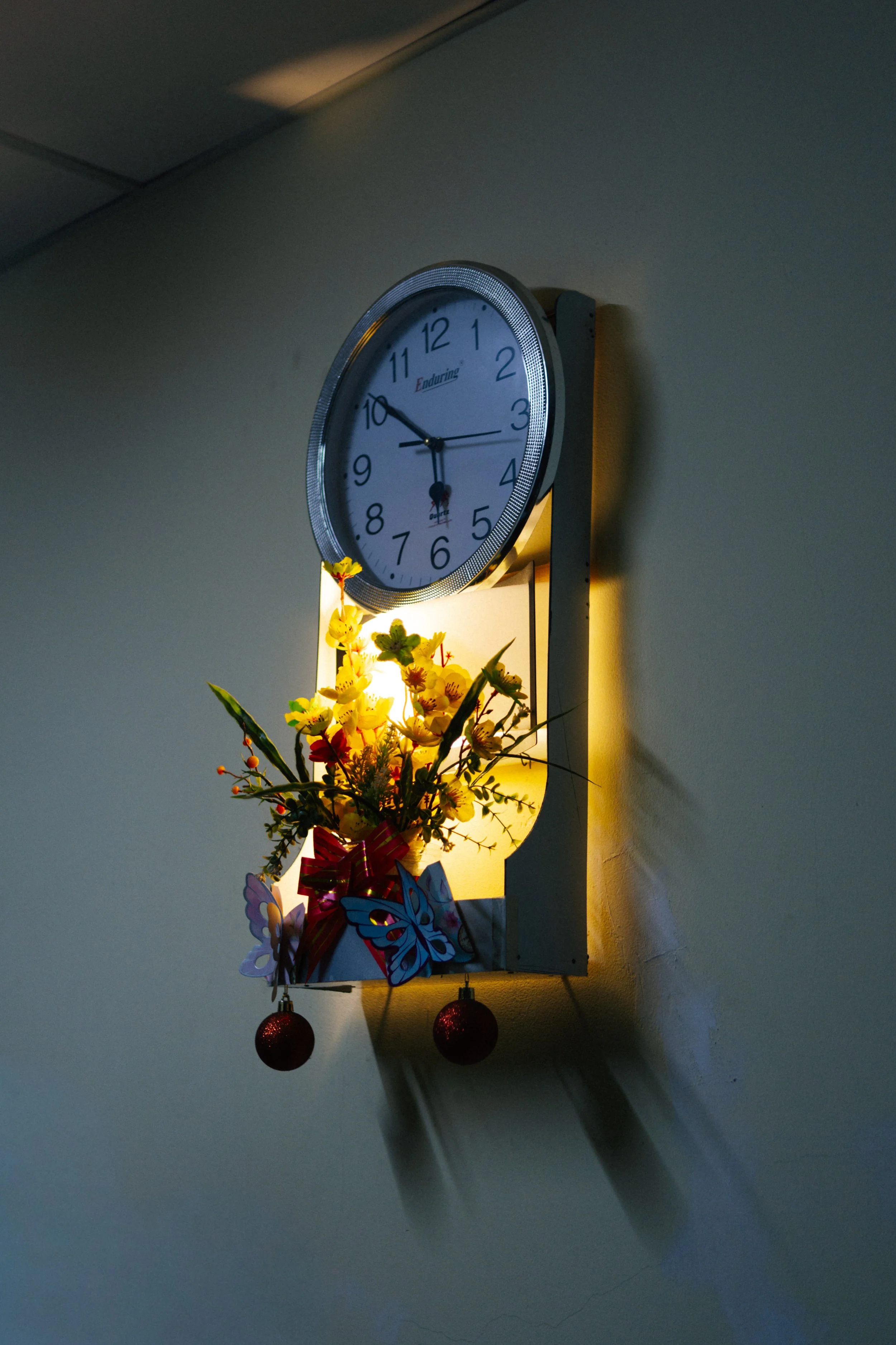 A round wall clock showing the time as 9:50, with a decorative flower arrangement and butterfly ornaments hanging below. The clock is mounted on a beige wall, illuminated by a light source.