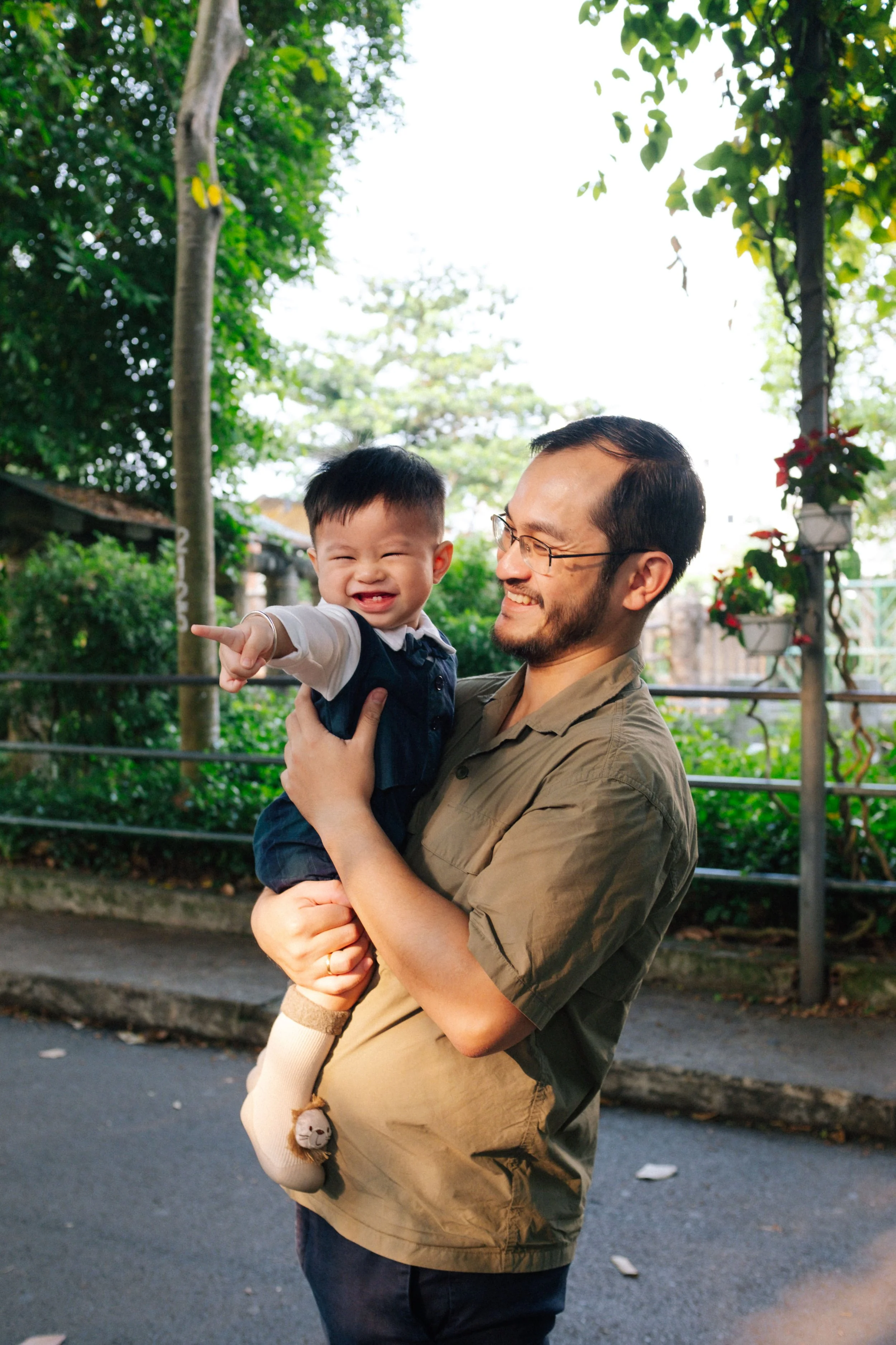 A man holding a smiling young boy outdoors, surrounded by trees and greenery.