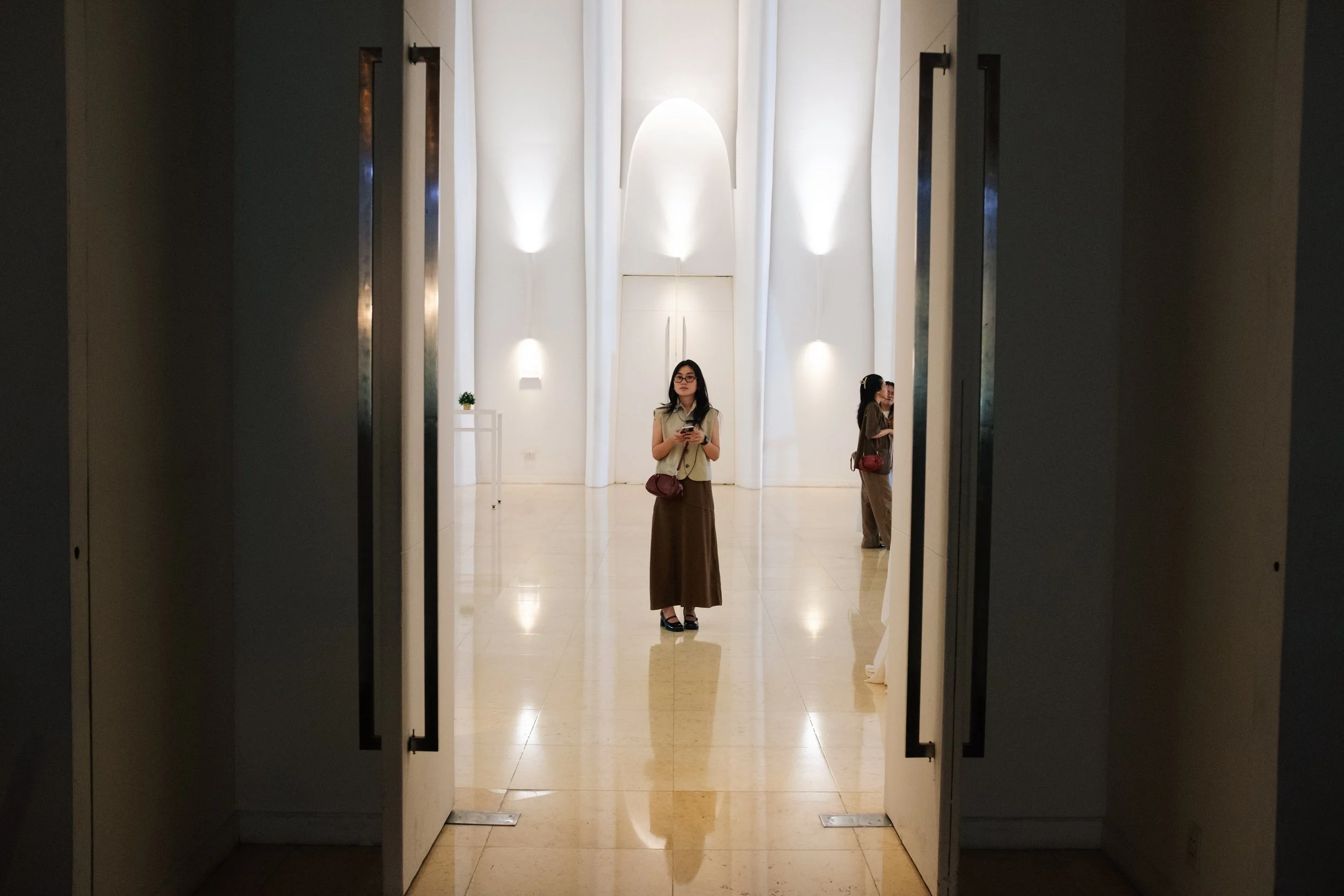 Woman in glasses standing inside an art gallery, looking at her phone, reflected in a mirror, with two other people in the background.