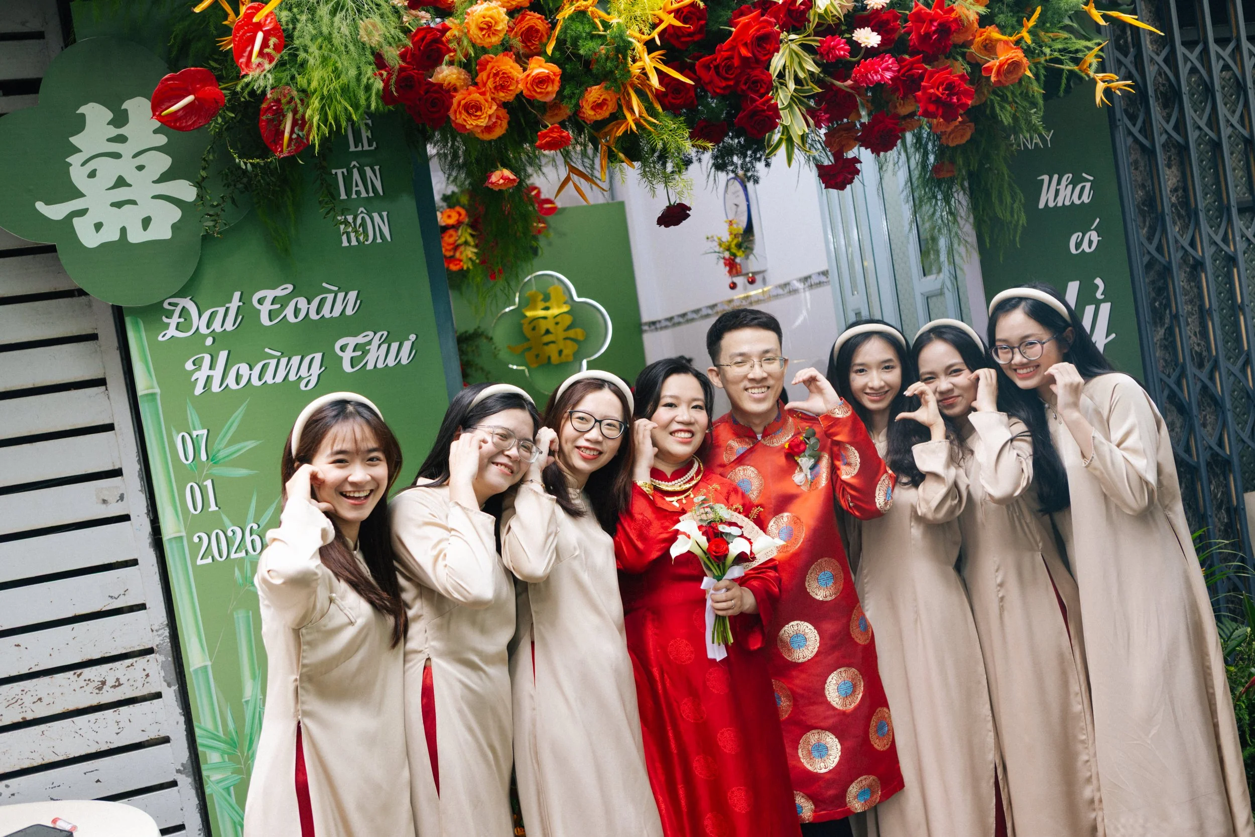 A group of people celebrating a wedding, with the bride and groom at the center wearing traditional Vietnamese wedding attire, surrounded by friends in beige dresses, posing in front of a green decorated backdrop with flowers and text in Vietnamese, 