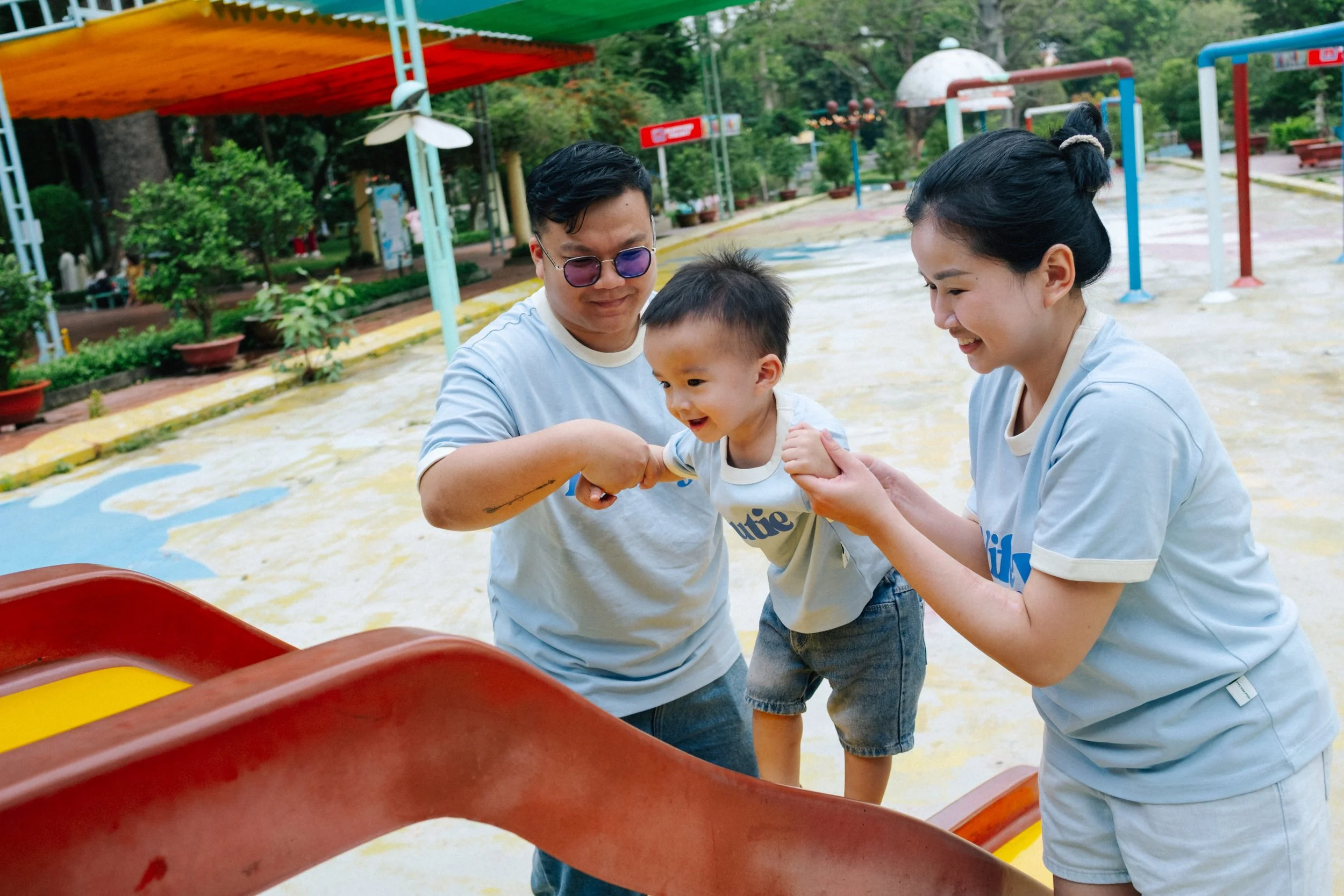 A young boy being helped by a man and woman at a playground, all smiling. The boy is mid-air on a slide, engaged in play.