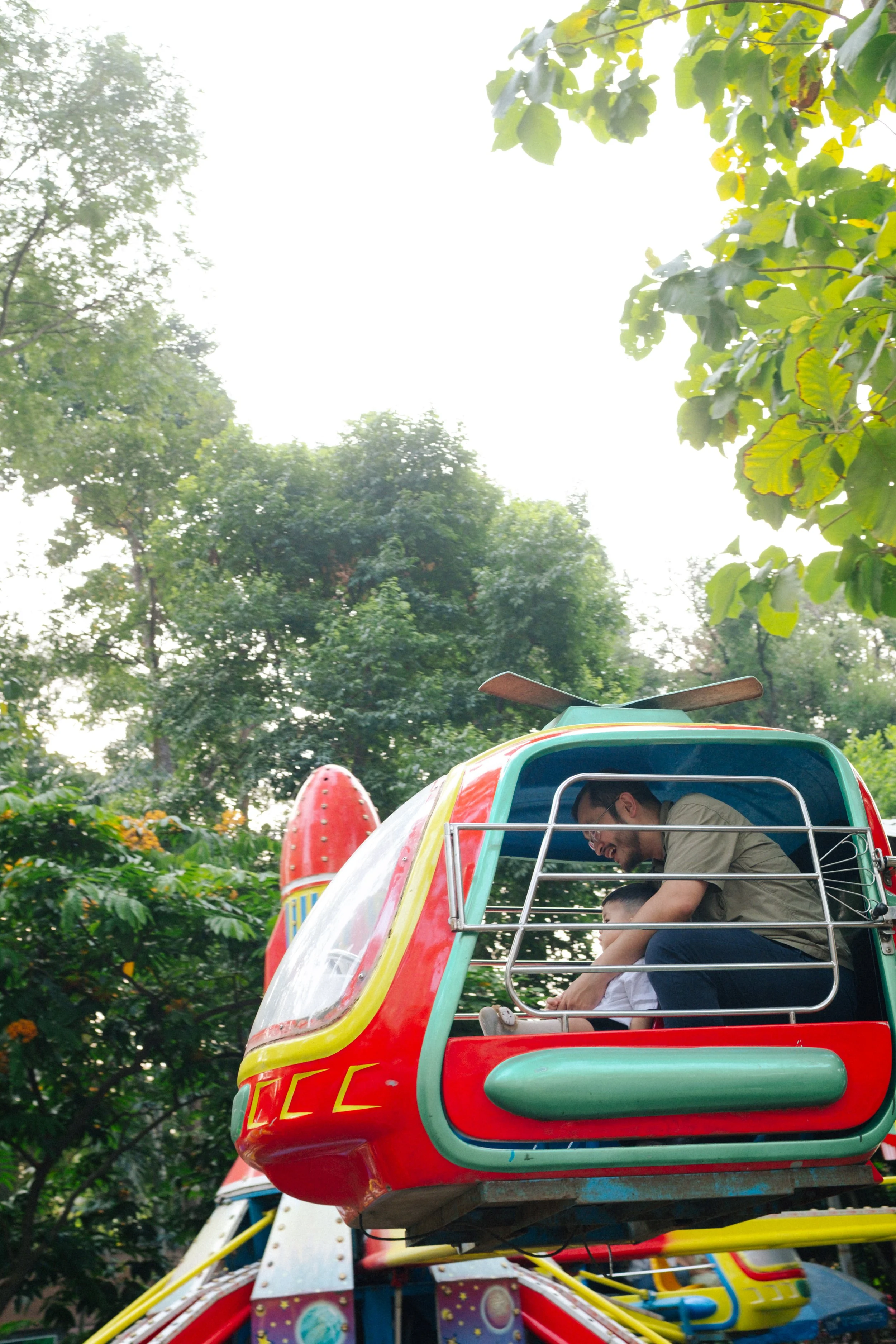 A man and a child riding in a small, colorful amusement ride shaped like a rocket in a park surrounded by greenery.
