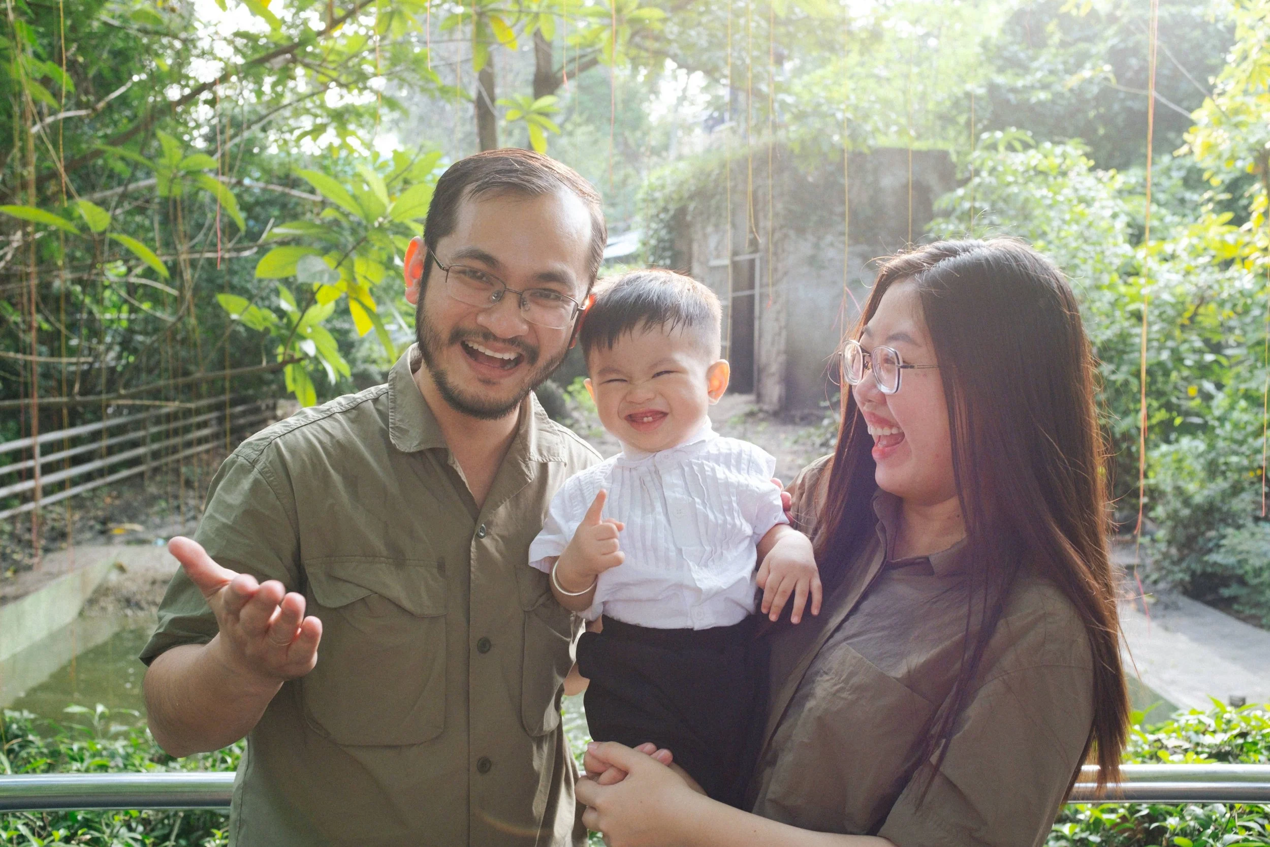 A happy family of three, including a man, a woman, and a baby, standing outdoors in a lush garden. The man and woman are smiling and looking at the baby, who is also smiling and pointing.