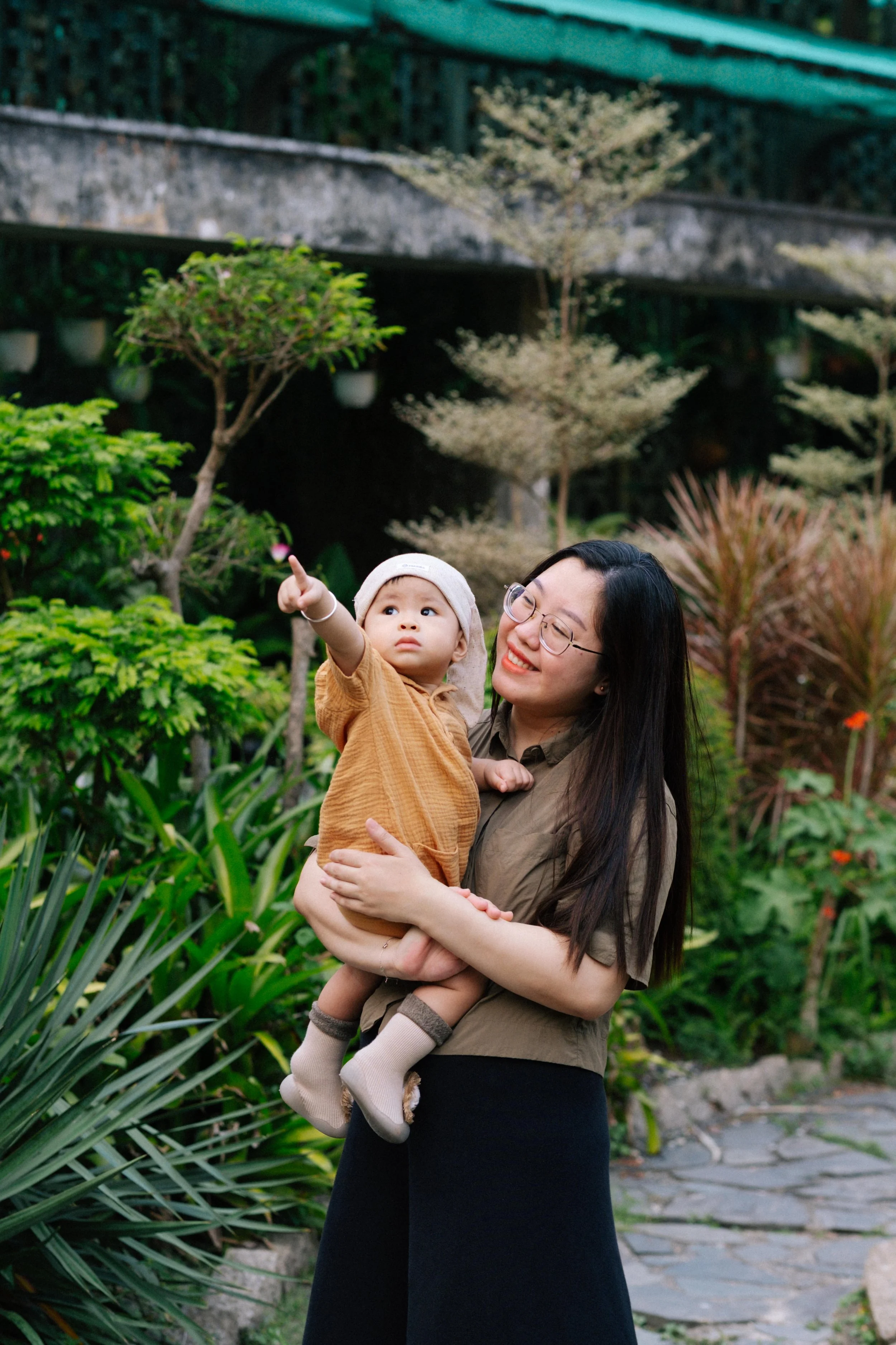 A woman in glasses holding a young child in a garden with greenery and trees, the child pointing at something in the sky.