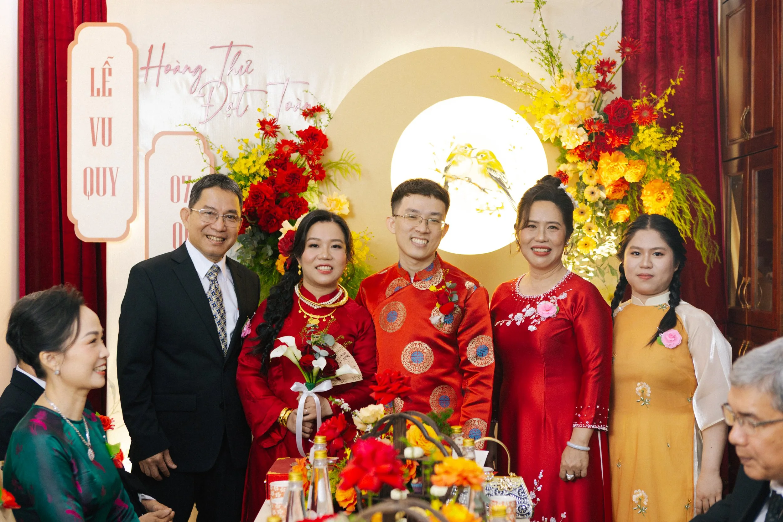 A Vietnamese wedding celebration with couples and family members dressed in traditional attire, standing in front of floral decorations and artwork featuring a bird and yellow flowers.