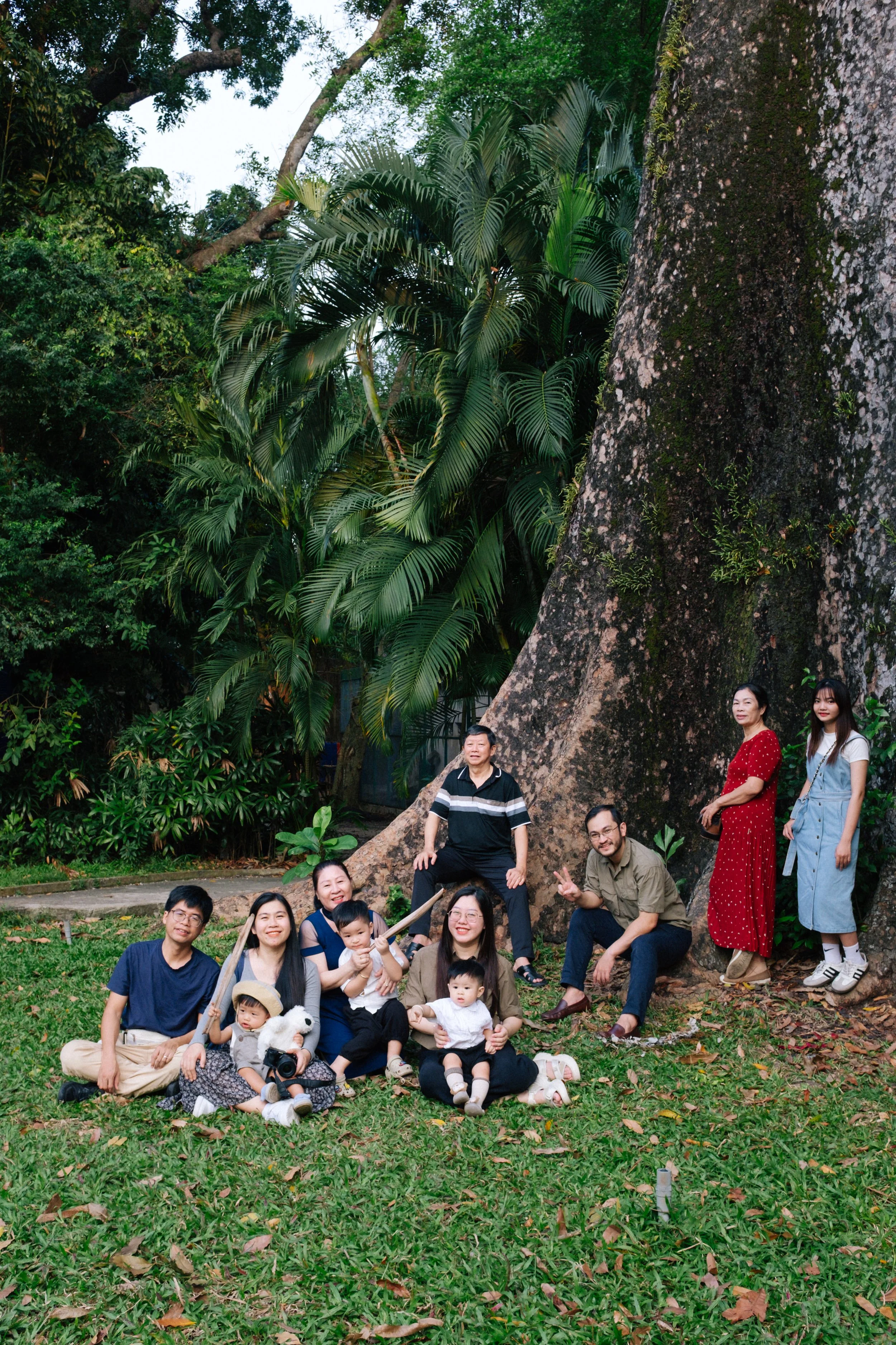 A group of ten people, including adults and children, are gathered outdoors in front of a large tree with green foliage. They are posing for a photo on the grass, with some sitting, standing, and kneeling, smiling and making peace signs. The backgrou