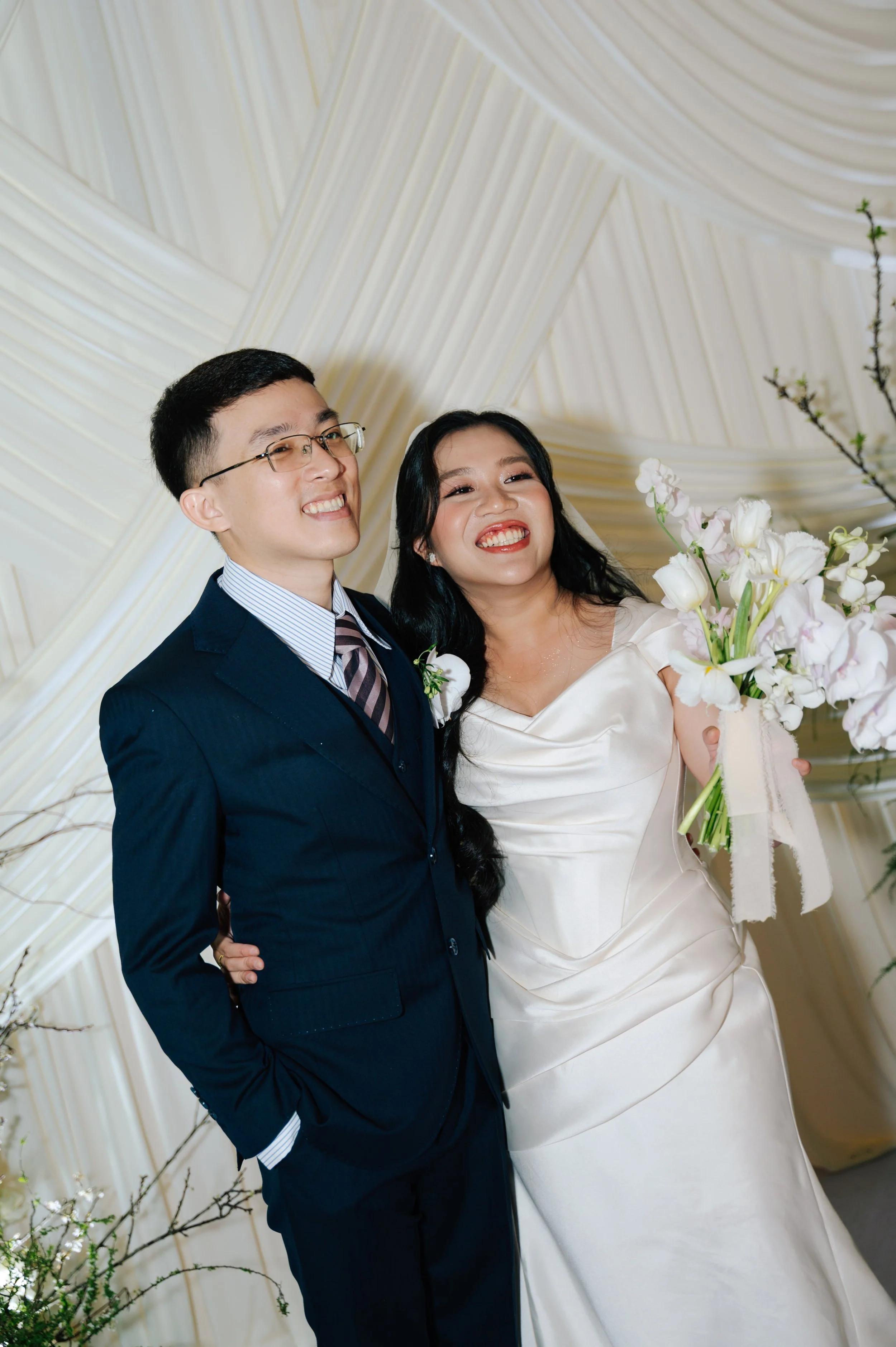 A bride and groom smiling at their wedding, standing in front of a cream-colored draped backdrop. The bride is holding a bouquet of white and pale pink flowers.