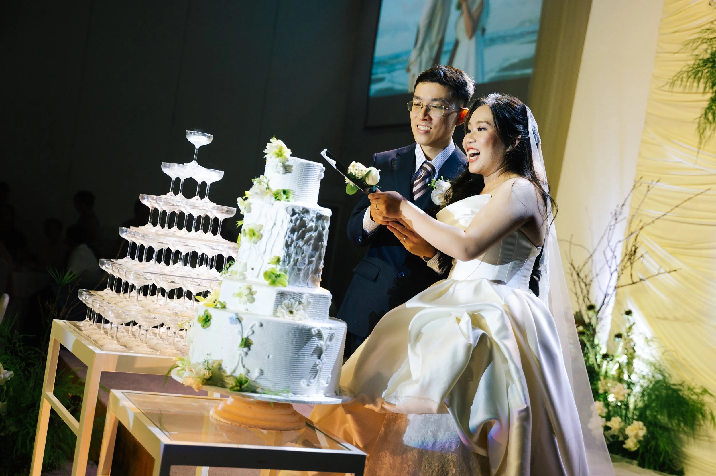 A couple in wedding attire cutting a wedding cake together, standing beside a tower of champagne glasses, during their wedding reception.