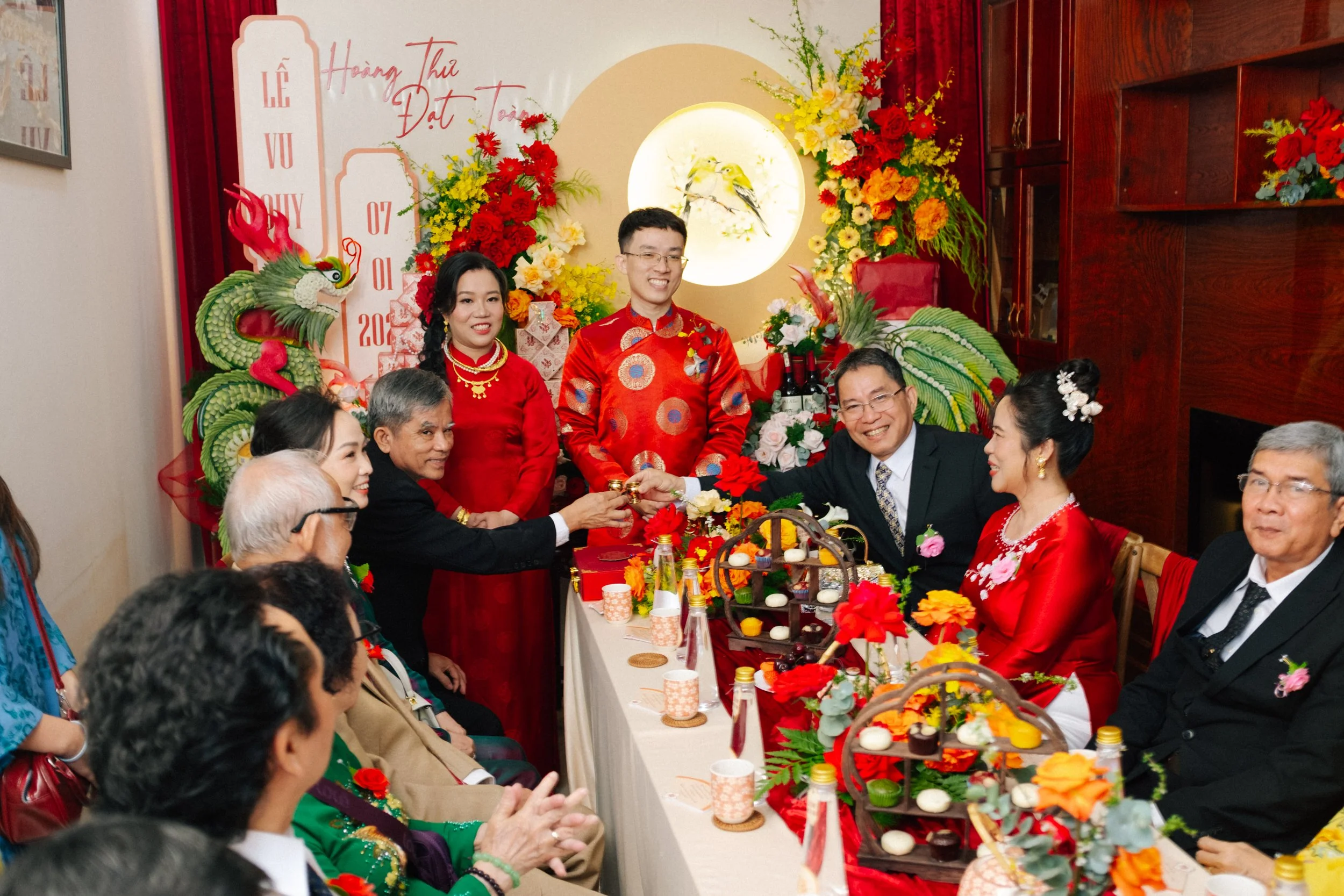 Family members celebrating a traditional Vietnamese wedding with red attire, floral decorations, and a dragon decoration in the background