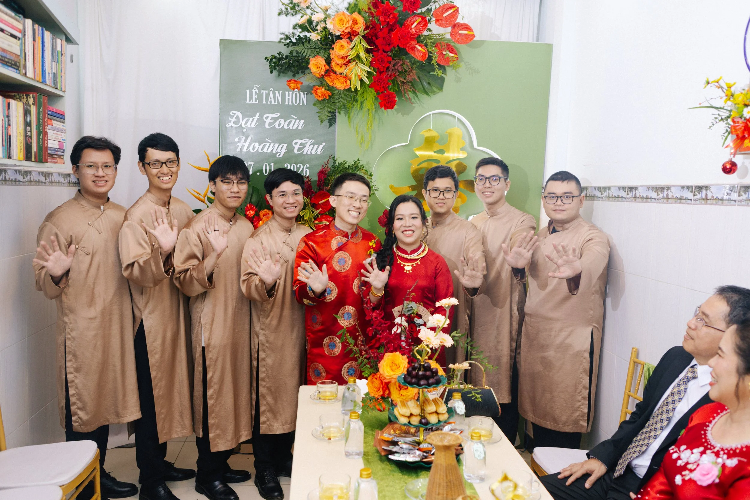 A group of nine young men and a woman in traditional Vietnamese attire, standing together at a wedding celebration, all smiling and waving. Two seated guests in formal attire are visible on the right. The background features a green board with Vietna