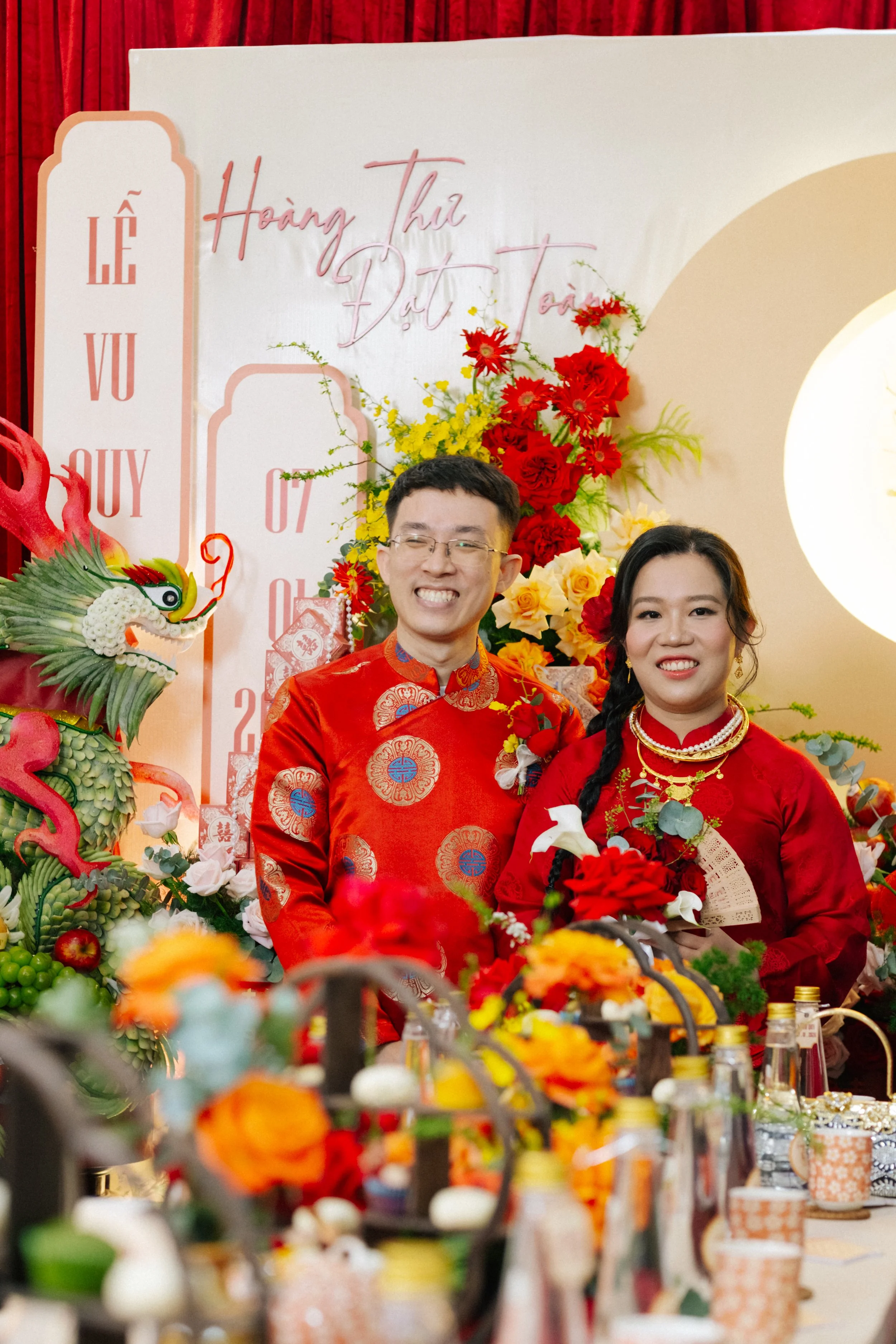 A Vietnamese couple dressed in traditional red attire celebrating a wedding, standing in front of a decorated backdrop with flowers and a dragon sculpture.