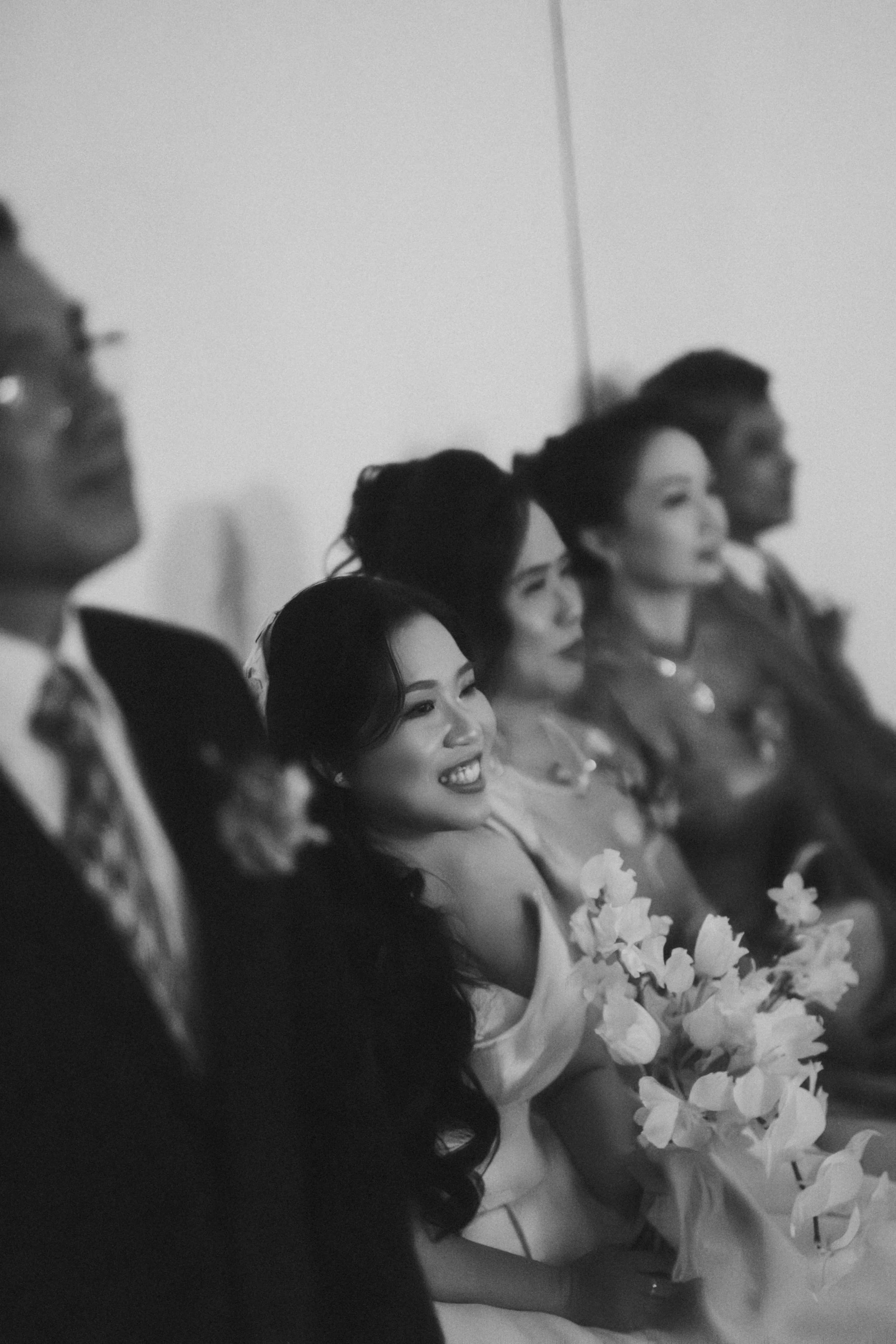 Black and white photo of five people sitting together at an event, smiling and watching something. One woman is holding a bouquet of flowers.
