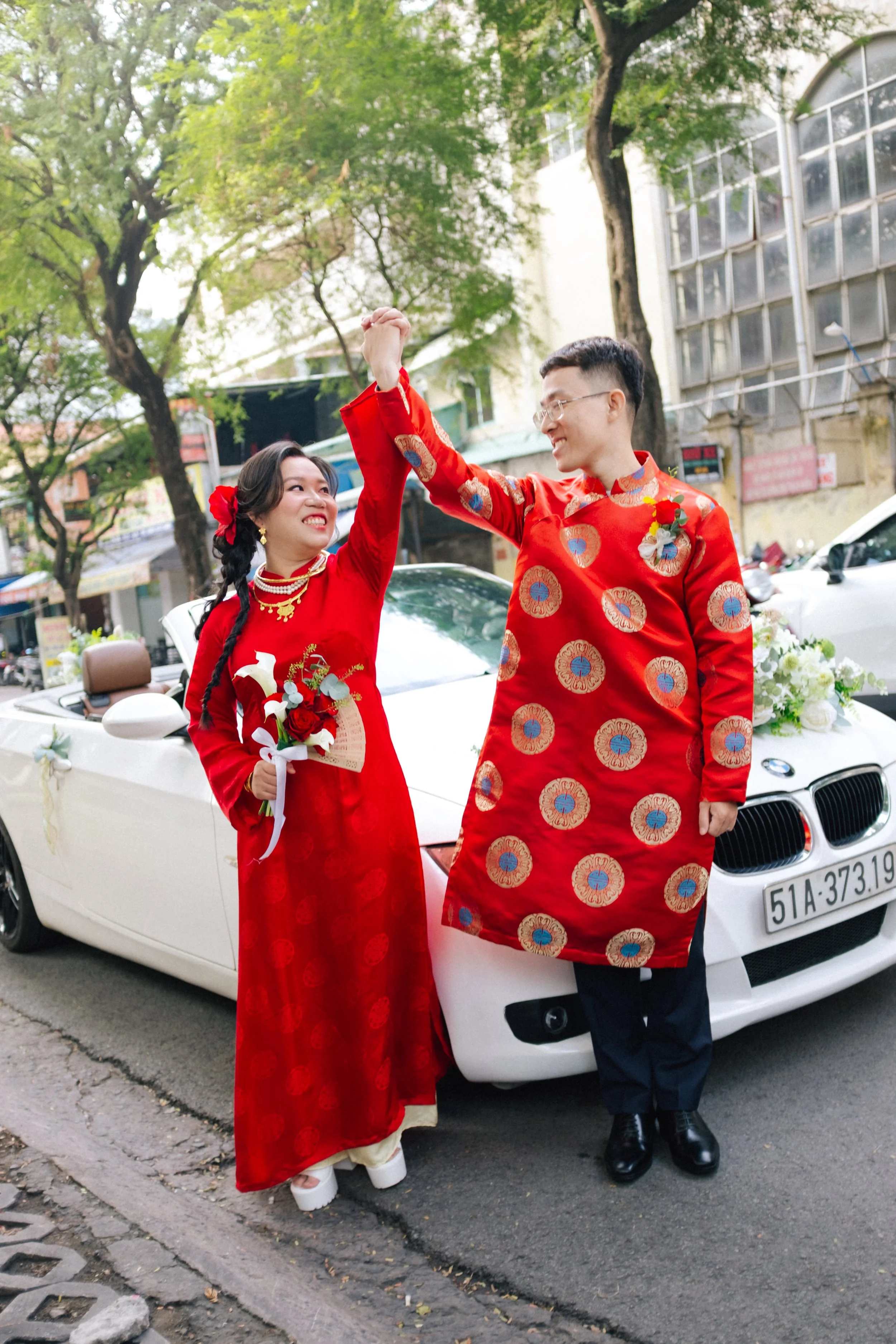 A couple dressed in traditional Vietnamese wedding attire, holding hands and smiling in front of a decorated white convertible car with flowers, on a street lined with trees.