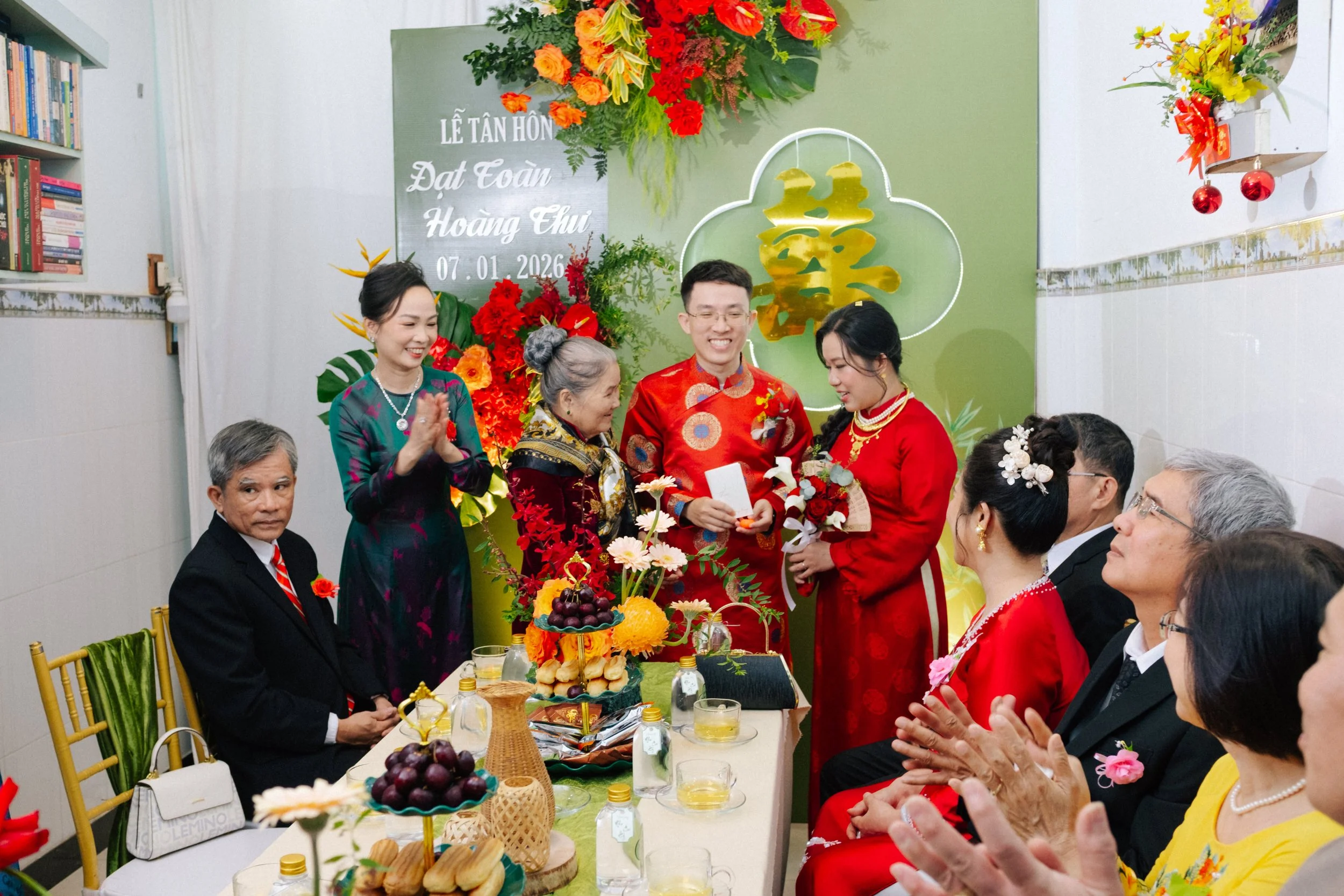 A Vietnamese wedding celebration with a bride and groom in red traditional clothing exchanging vows, surrounded by family and friends indoors, decorated with flowers and a green backdrop.