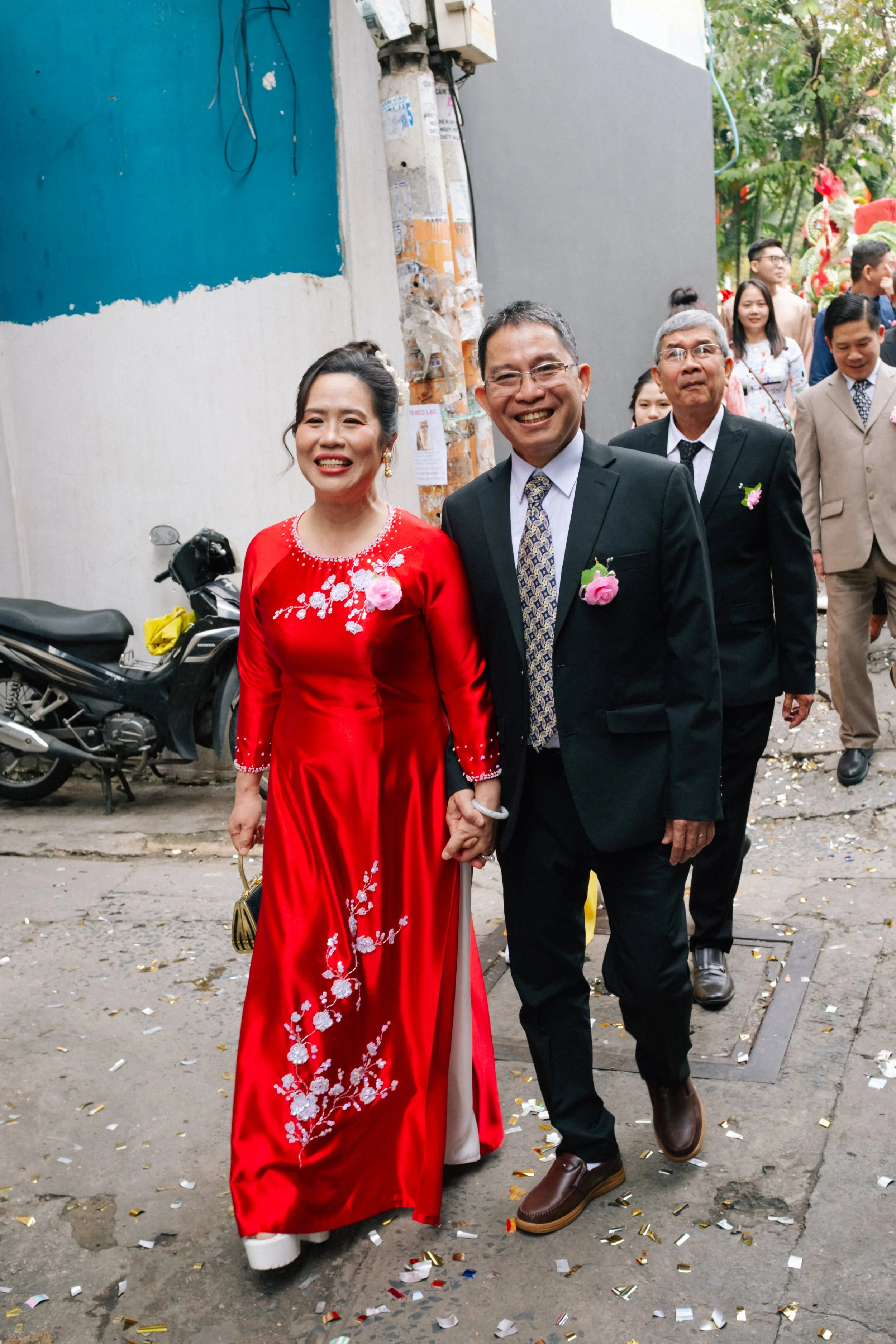 A joyful couple holding hands and smiling, dressed in traditional and formal attire, walking through a decorated street during a celebration or wedding, with other well-dressed guests in the background and colorful decorations including confetti on t