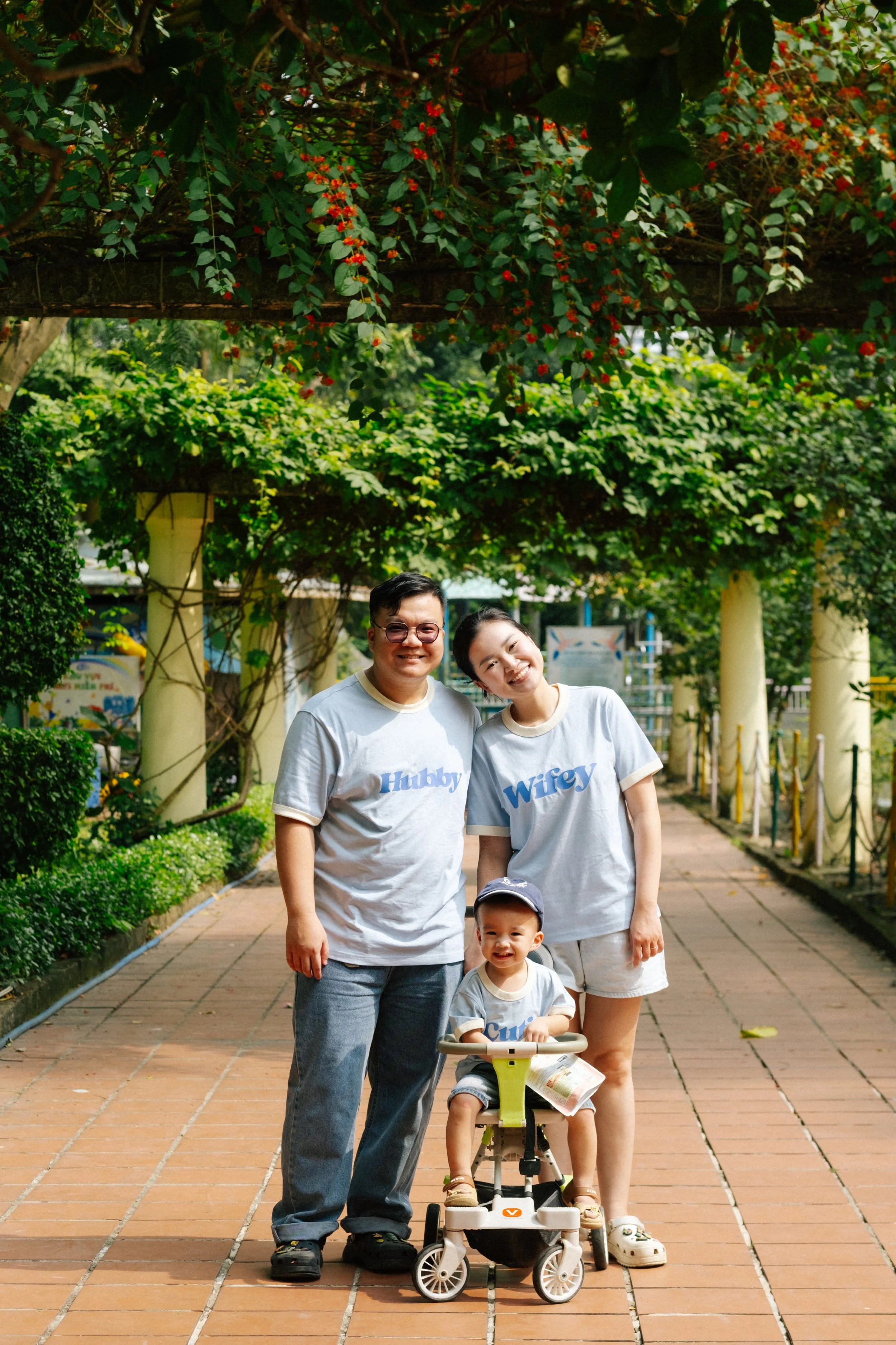 A family of three with matching gray t-shirts, saying Hubby, Wifey, and Cuter, standing together outdoors on a paved path in a park with greenery and trees around them. The young child is sitting in a small stroller, smiling, with a cap and a magazin