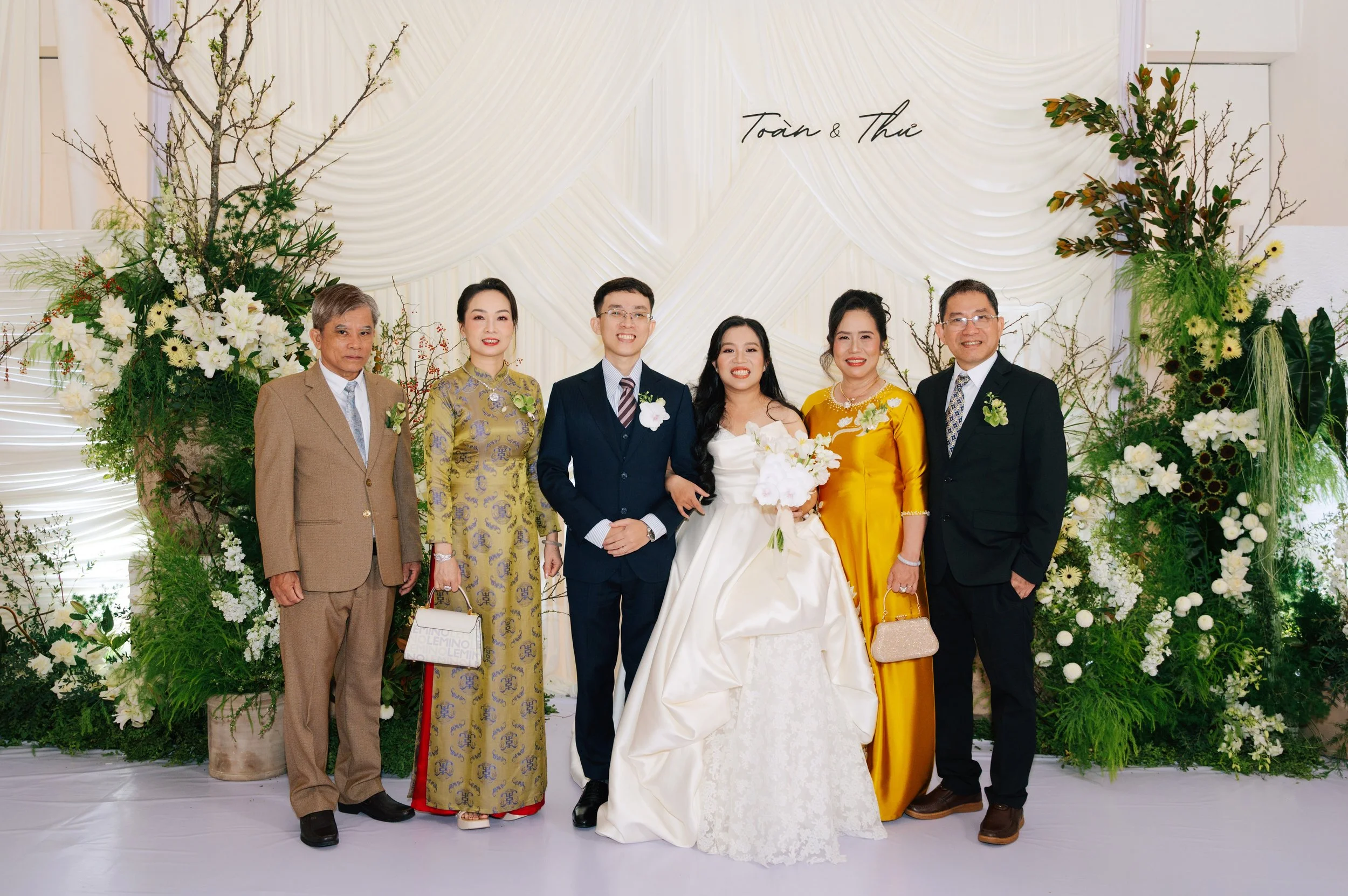 Group of seven people, including bride and groom, posing at a wedding reception with floral backdrop and guests dressed formally.