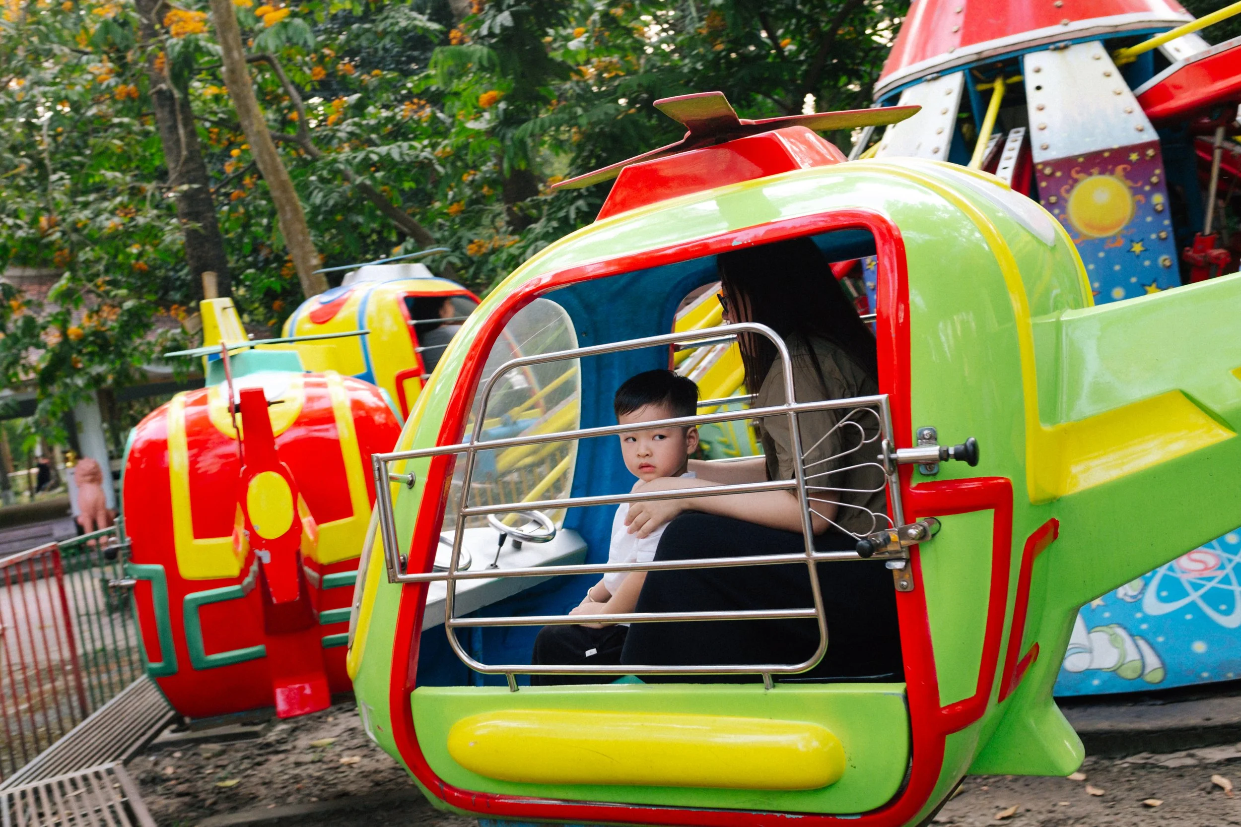 A young boy and a woman sitting inside a colorful helicopter ride at an amusement park.