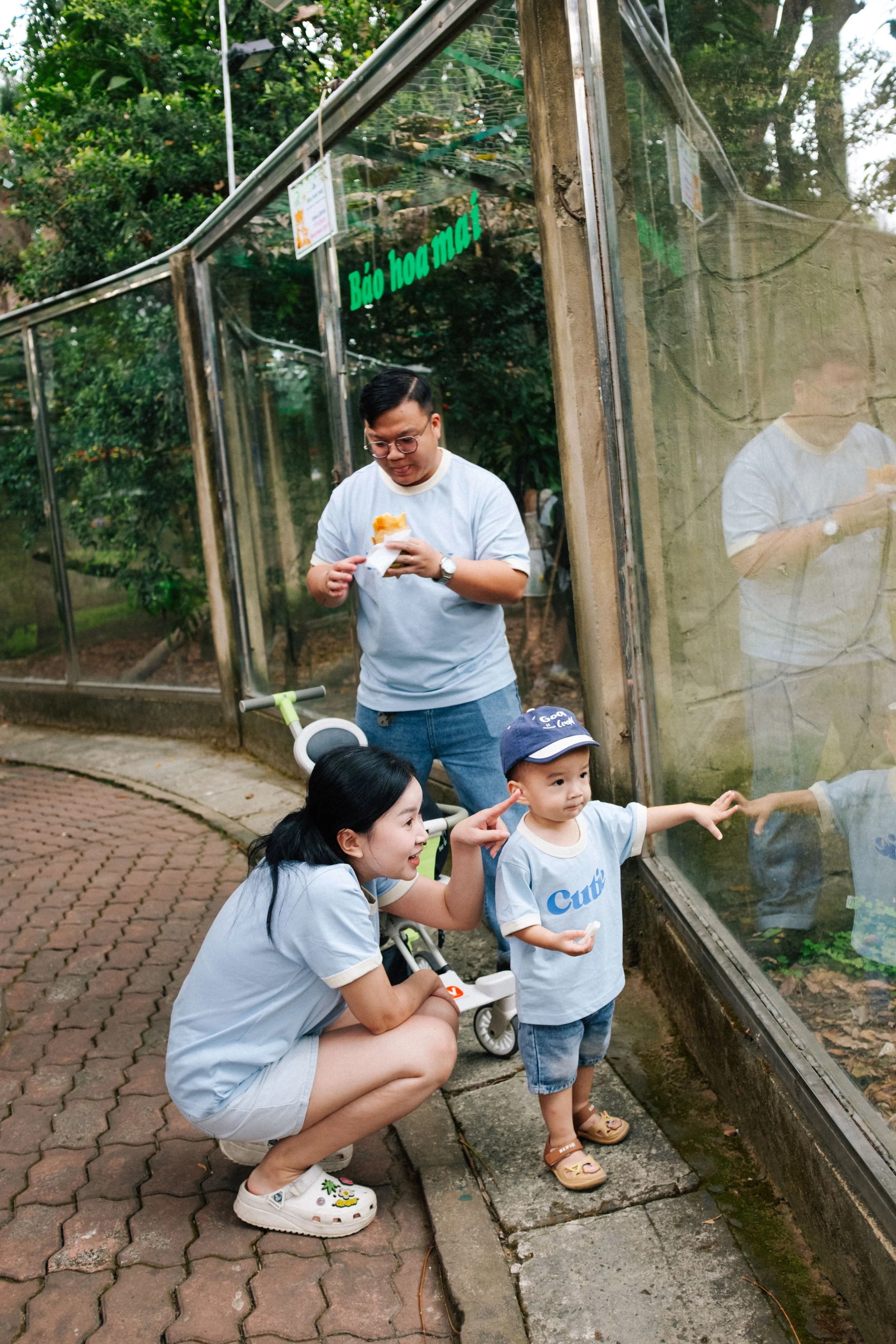 A family of three at a zoo, observing animals through a glass enclosure. The woman is crouching, pointing at the animals, a young boy beside her is pointing and wearing a Build cap, and the man holds a snack, standing behind them. There is a stroller