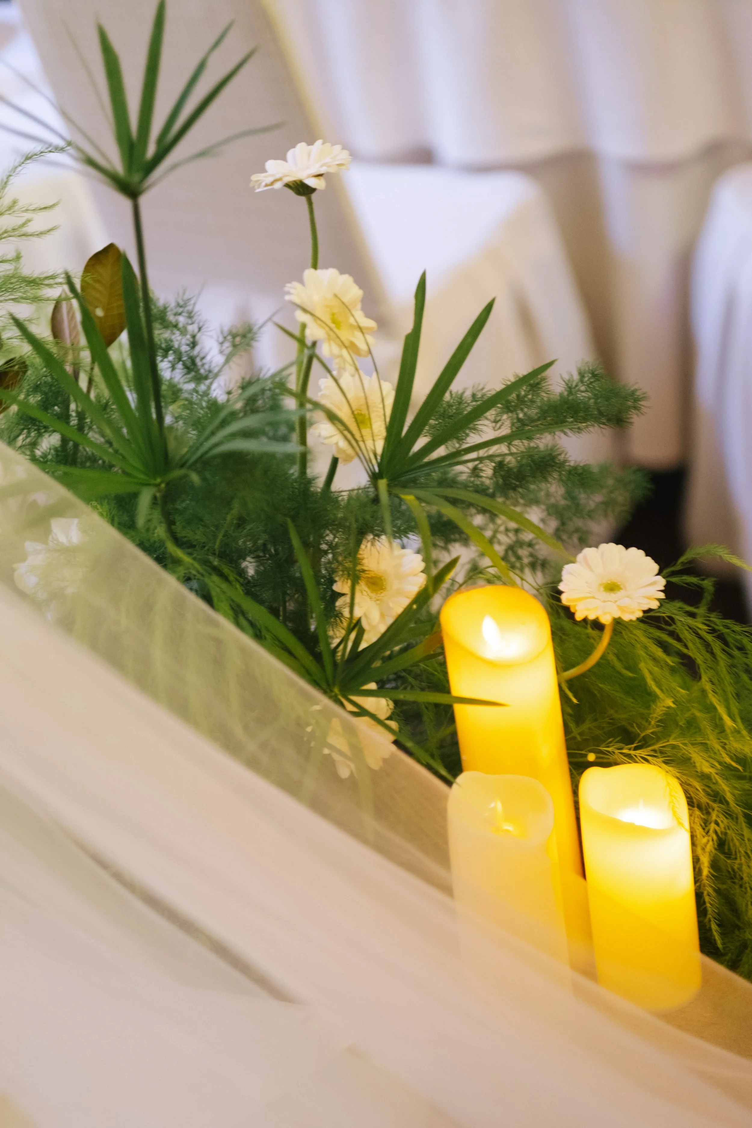 A floral arrangement with white flowers, green leaves, and candles on a table