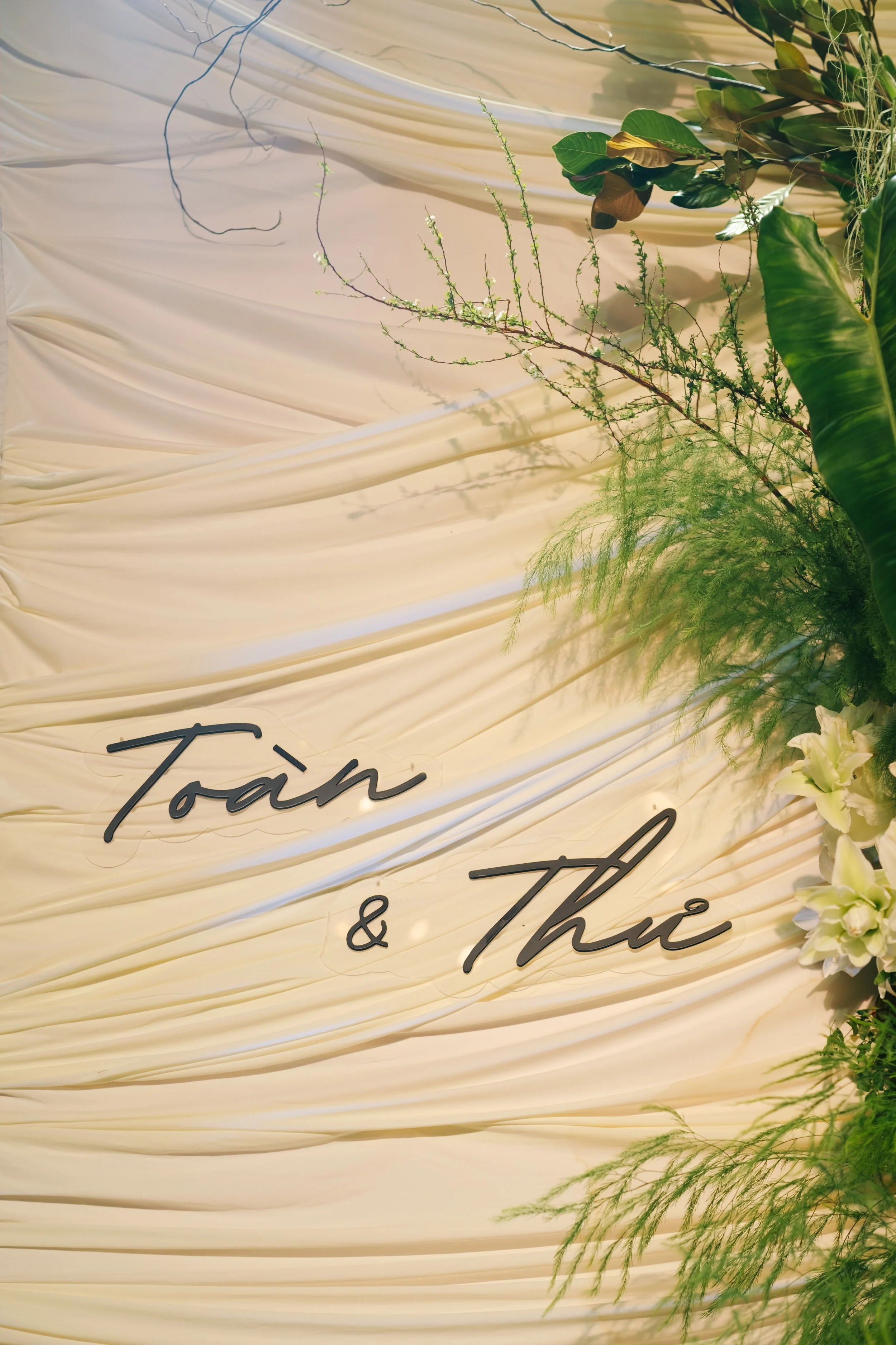 Decorative floral backdrop with cream-colored draped fabric featuring a black neon sign that reads 'Toàn & Thu', decorated with lush green leaves and white flowers.