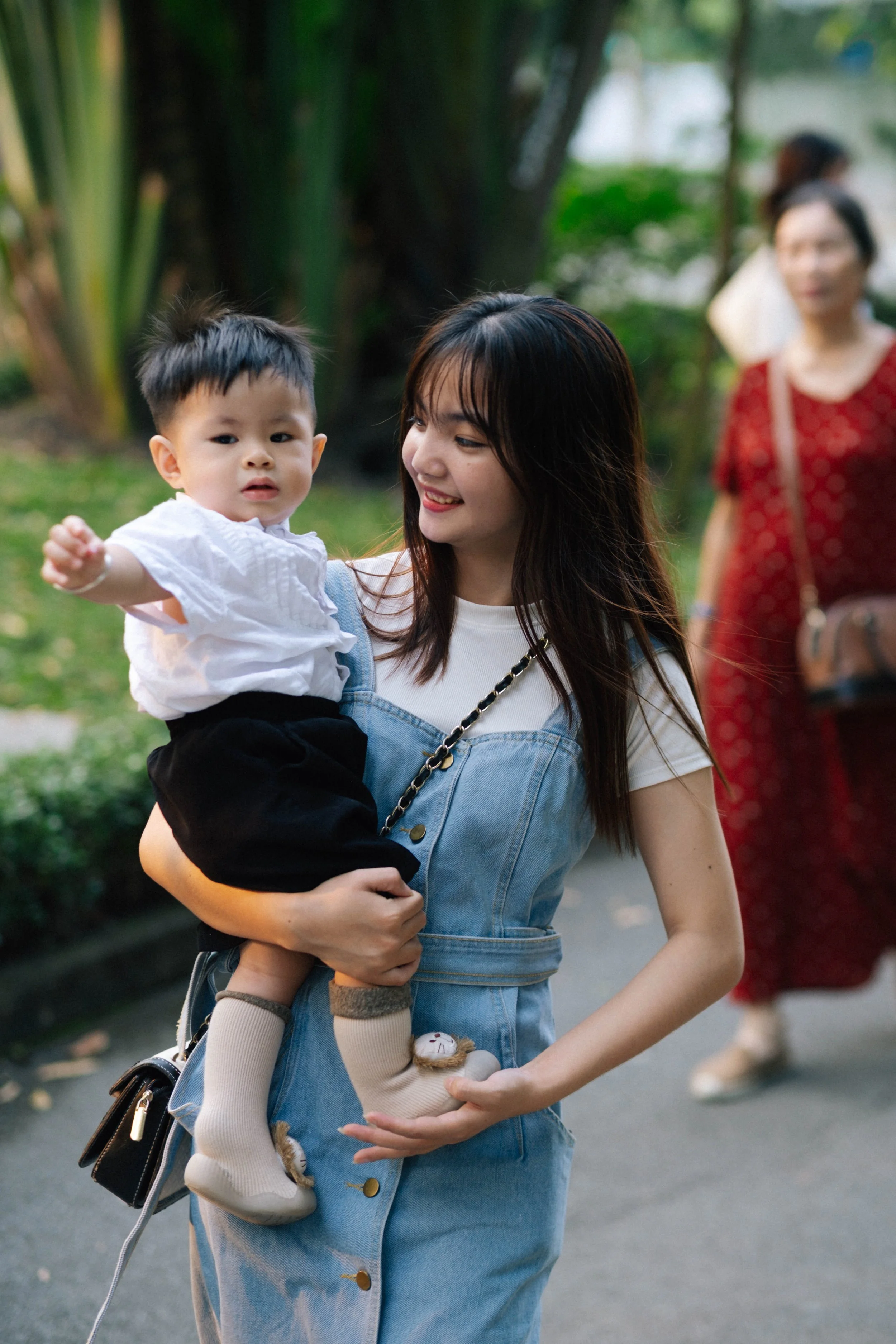 A young woman holding a small child in an outdoor setting, with another woman walking in the background.