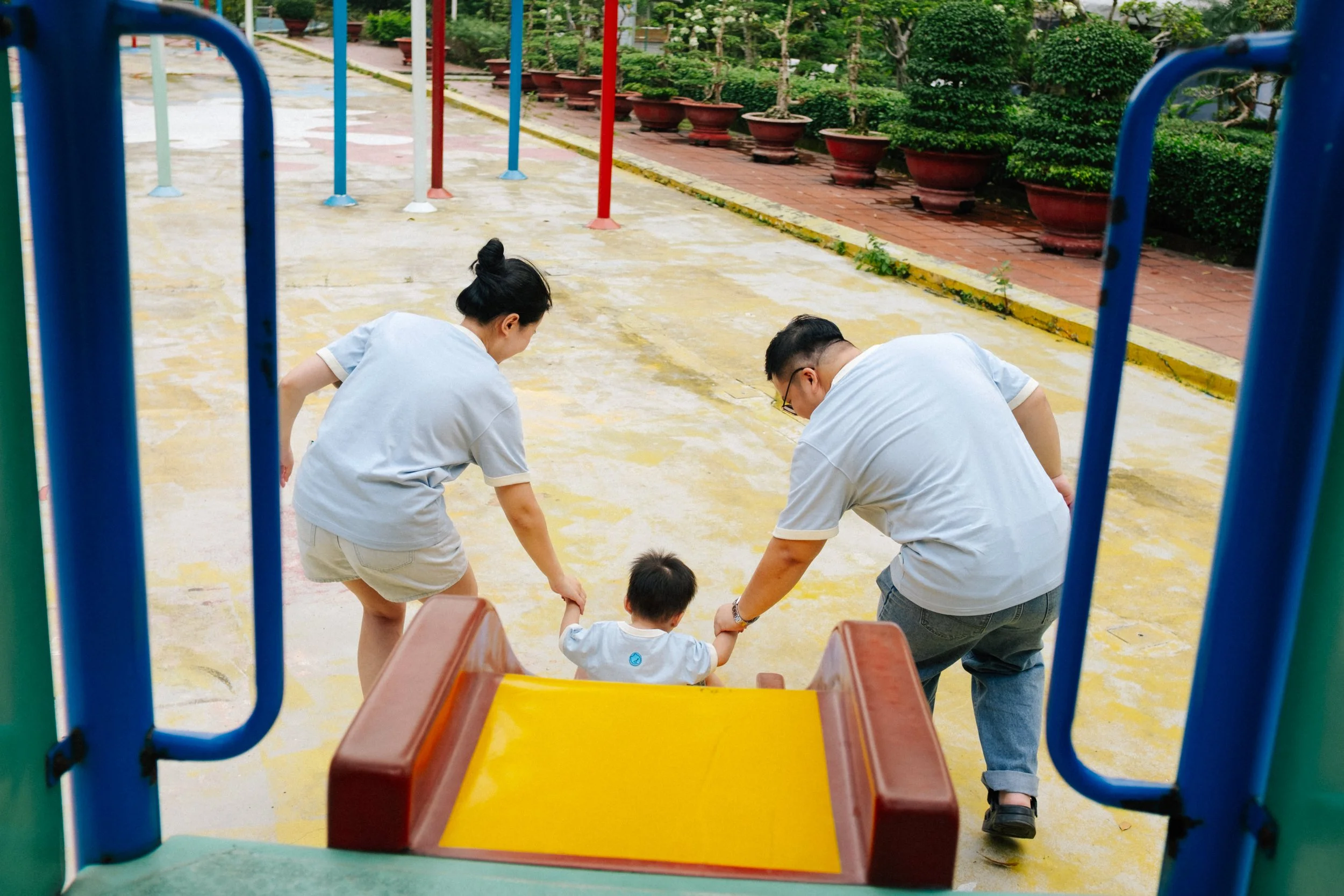 A young child sitting on a small yellow slide at a playground, holding hands with two adults, possibly the child's parents, as they help the child climb up the slide. The playground has a wet surface and is surrounded by potted plants and greenery.